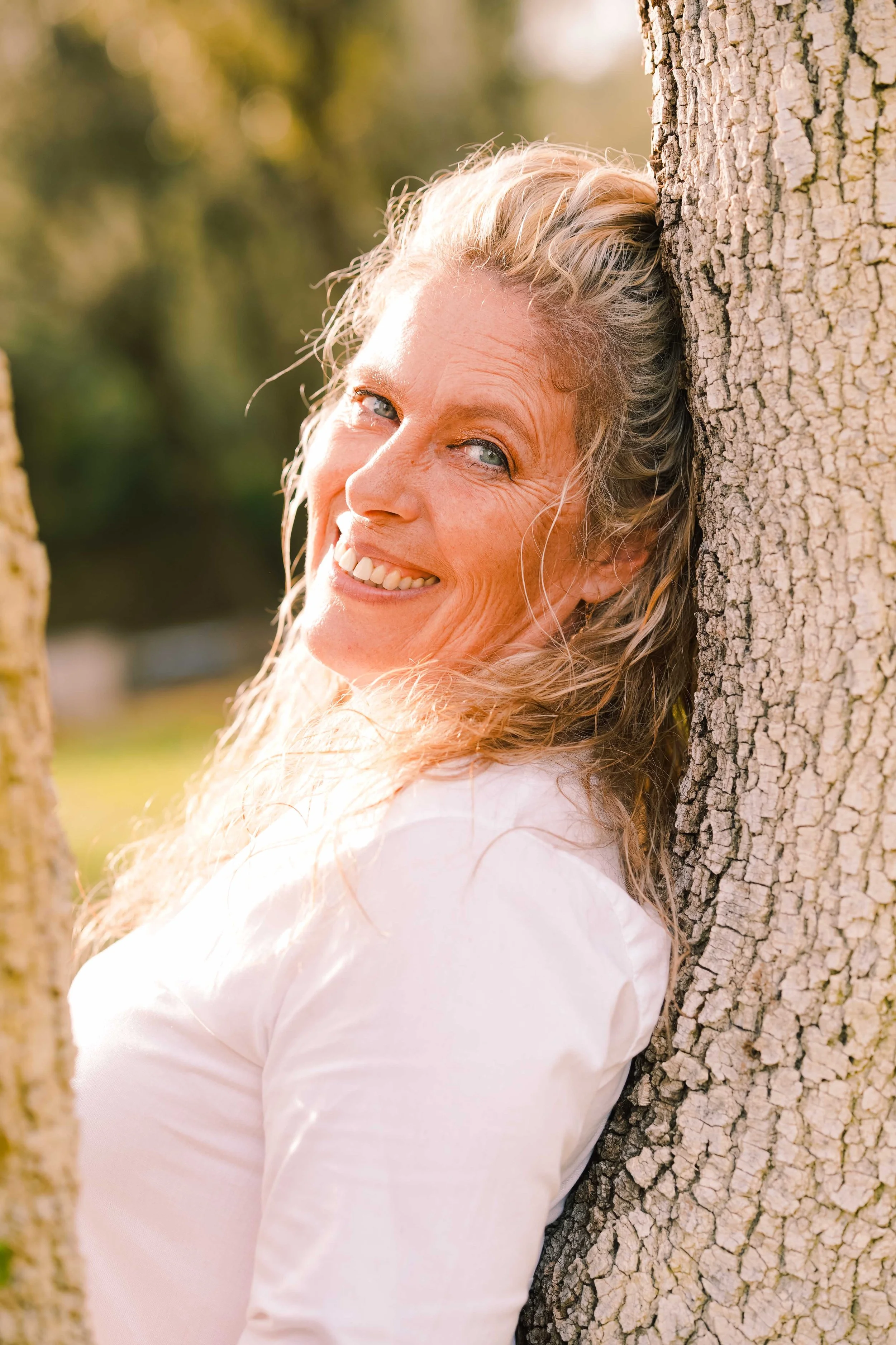 Femme souriante appuyée contre un arbre en plein air.
