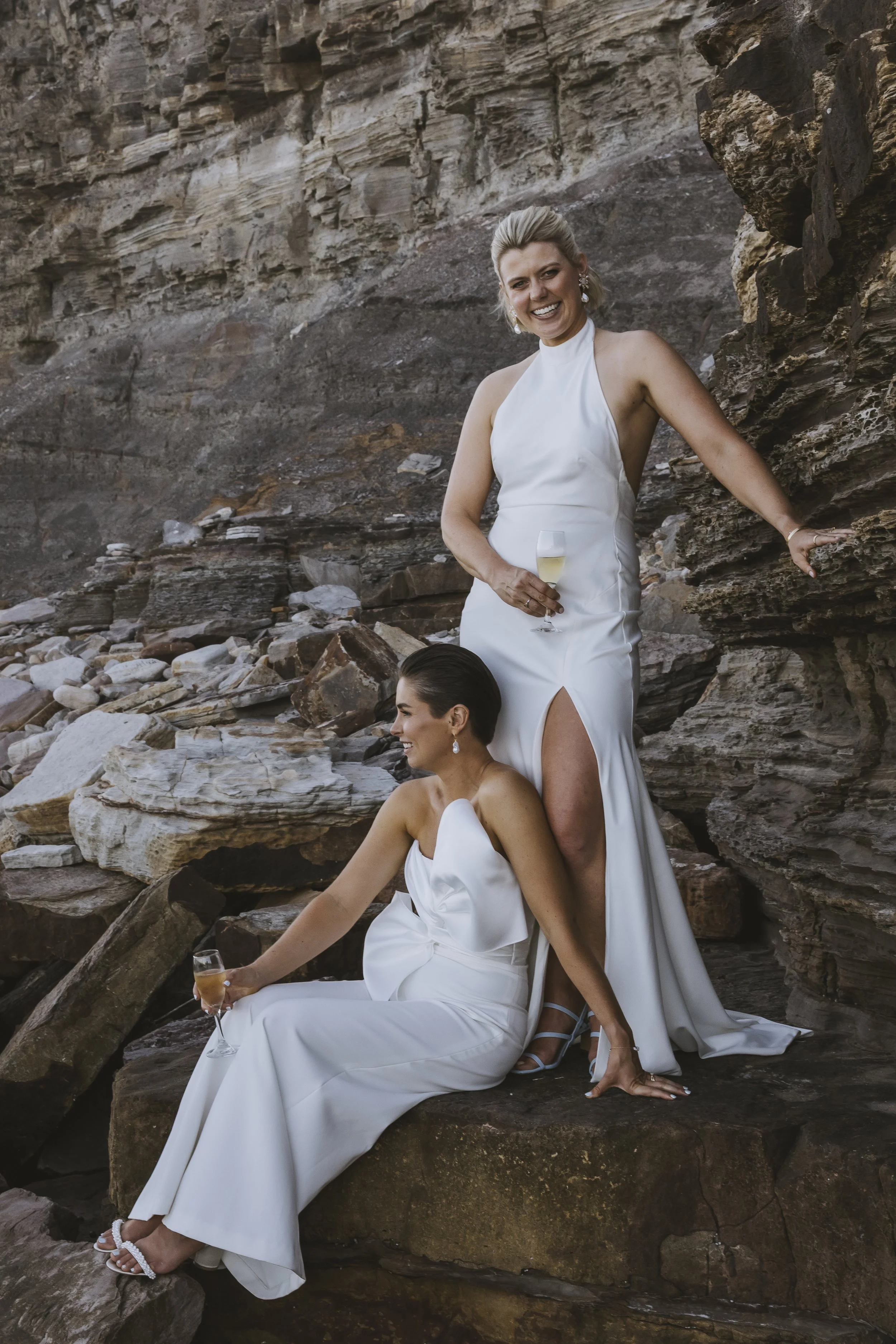 Two women in white dresses at a rocky outdoor location, one standing and holding a glass of champagne, the other sitting on a rock also holding champagne, both smiling.