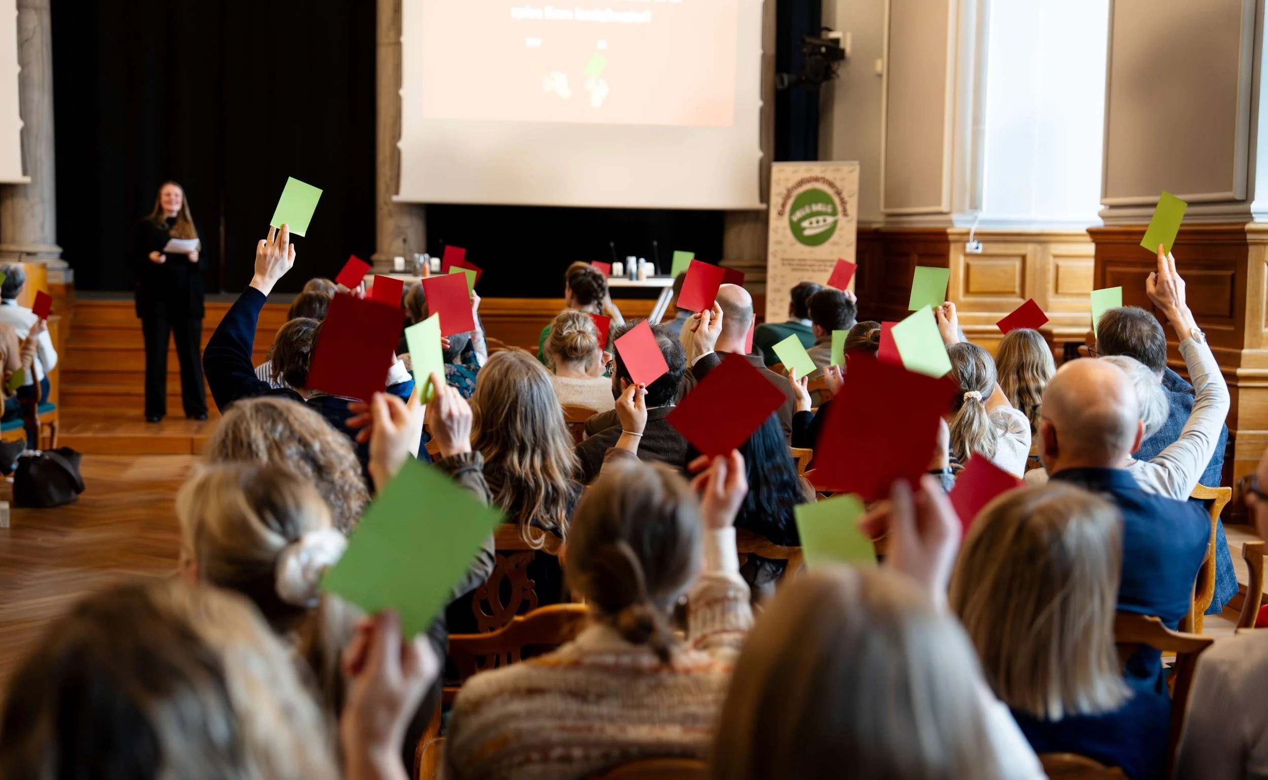 En stor forsamling af mennesker deltager i en stemmeafstemning med røde og grønne stemmesedler i en konference sal, hvor en kvinde står foran snakker til publikum. Der er en stor skærm og en plankeværk med et logo i baggrunden.
