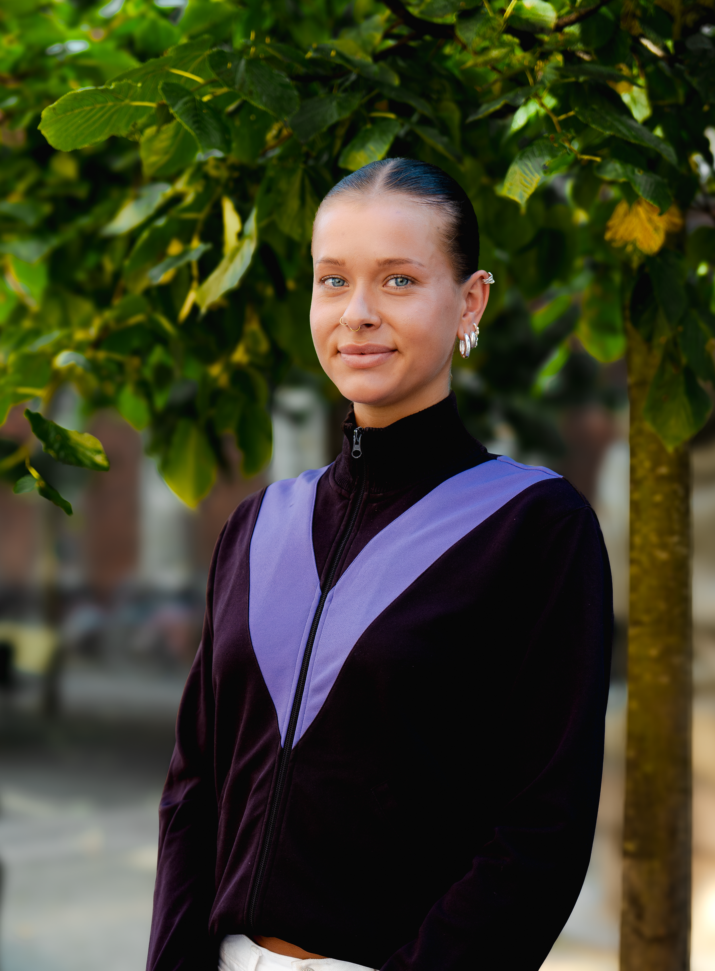 En ung kvinde med blå øjne, sort hår trukket tilbage, bærer store øreringe og en guldsmykke i øret, iført en sort og lilla trøje, står foran grønne blade.