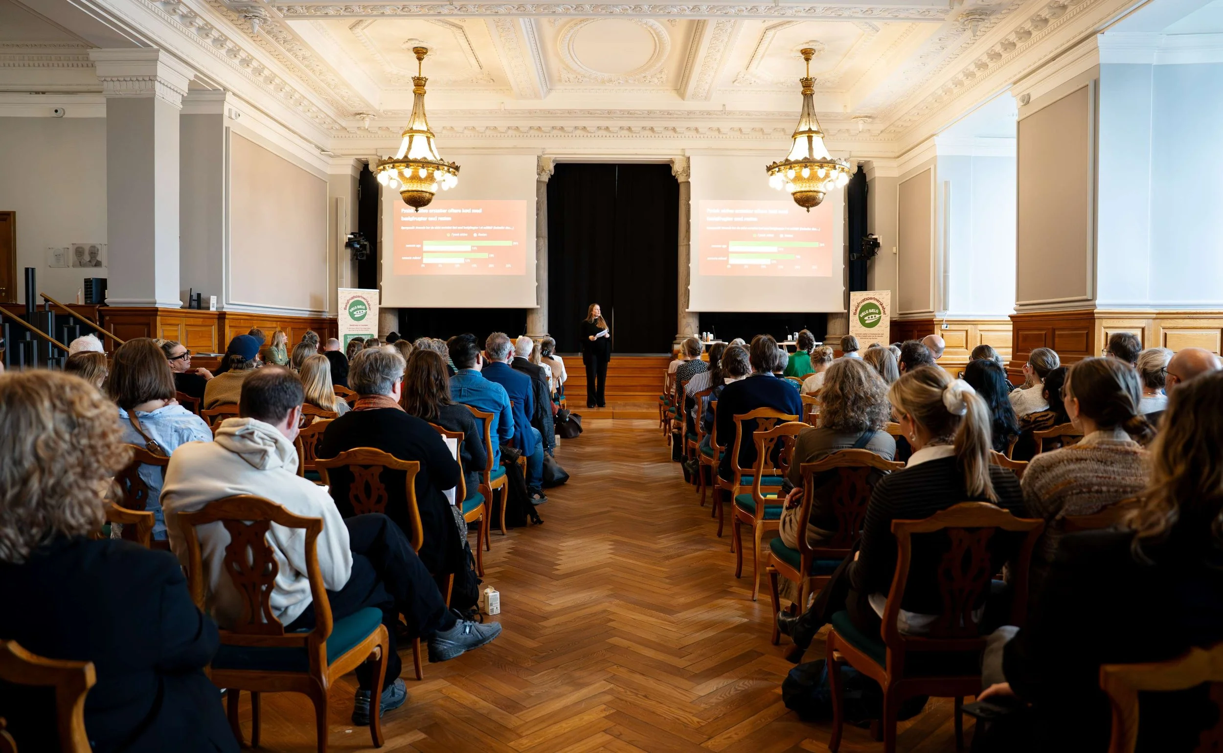 En stor forsamling mennesker sidder i et auditorie og lytter til en kvindelig taler foran to store skærme, der viser præsentationsslides. Rummet har høje lofter og smukke lysekroner.
