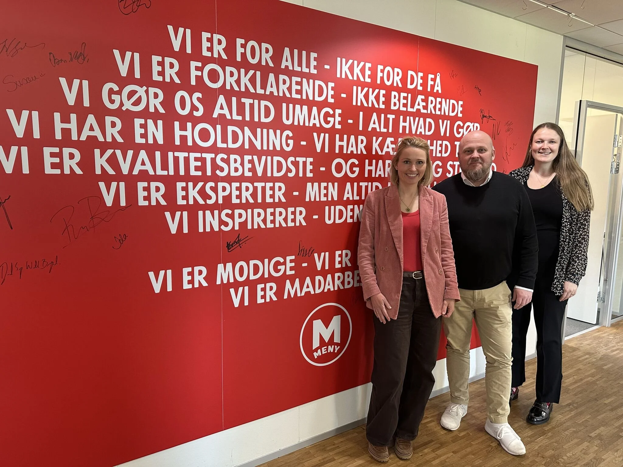 Tre mennesker står foran en rød væg med tekst, hvor der er skrevet flere autografer. De smiler og ser glade ud.