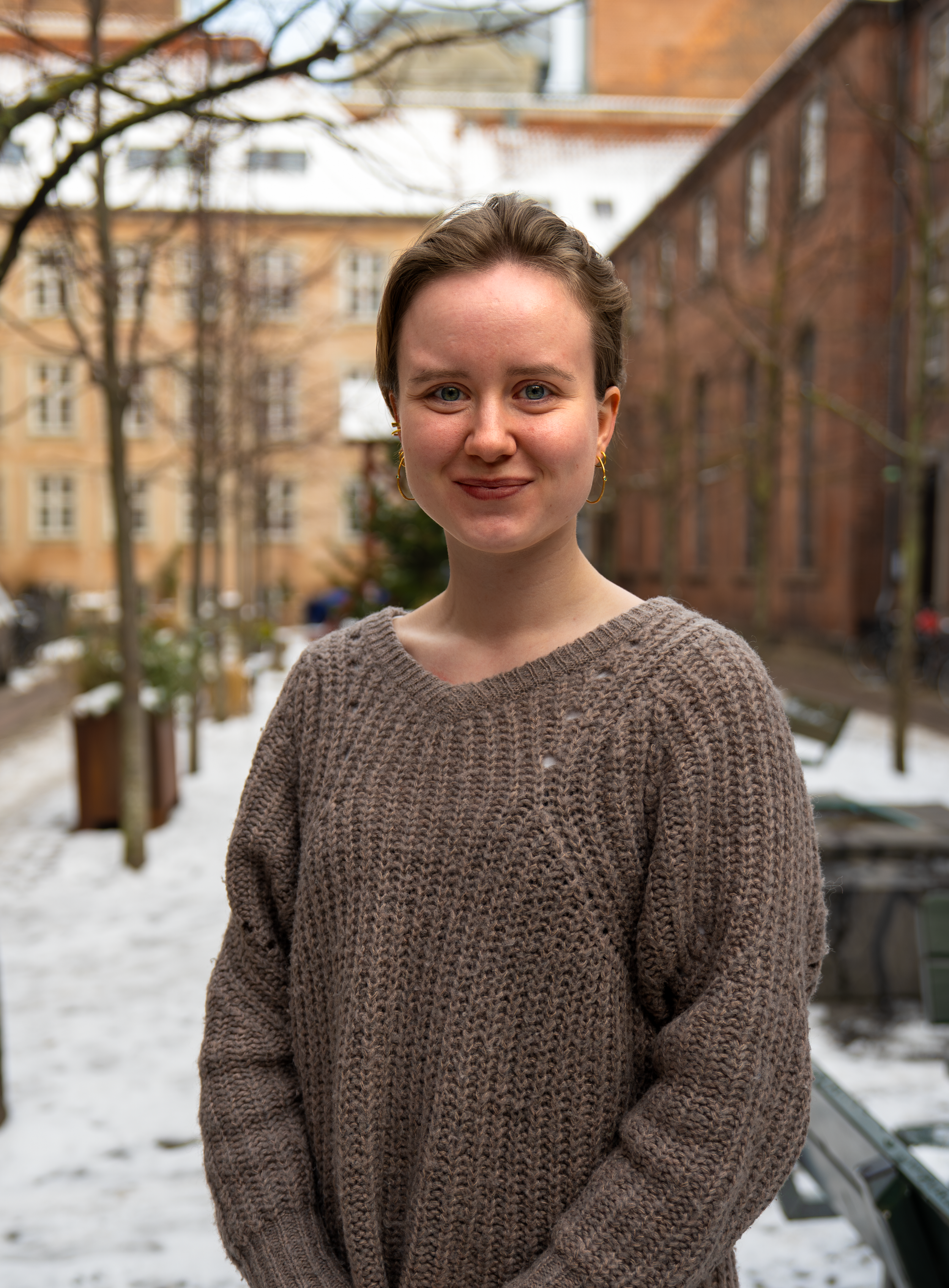 En ung kvinde med kort brunt hår og store guldørestikker står udenfor med sne på jorden og i baggrunden et træ og en bygning, iført en beige strikket sweater.
