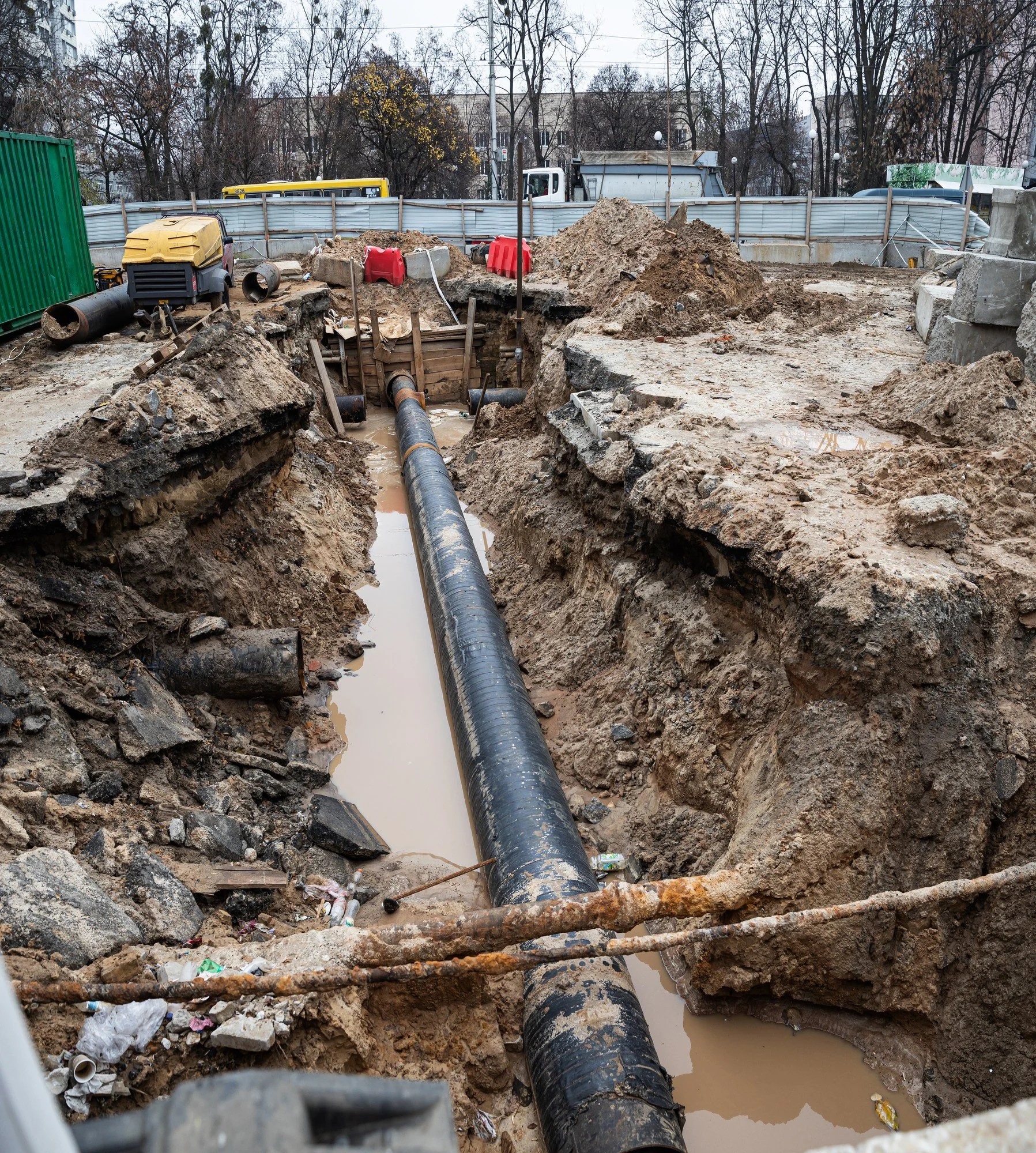 Construction site with trenches, large black pipe, dirt piles, and construction equipment surrounded by a temporary fence, with leafless trees and buildings in the background.