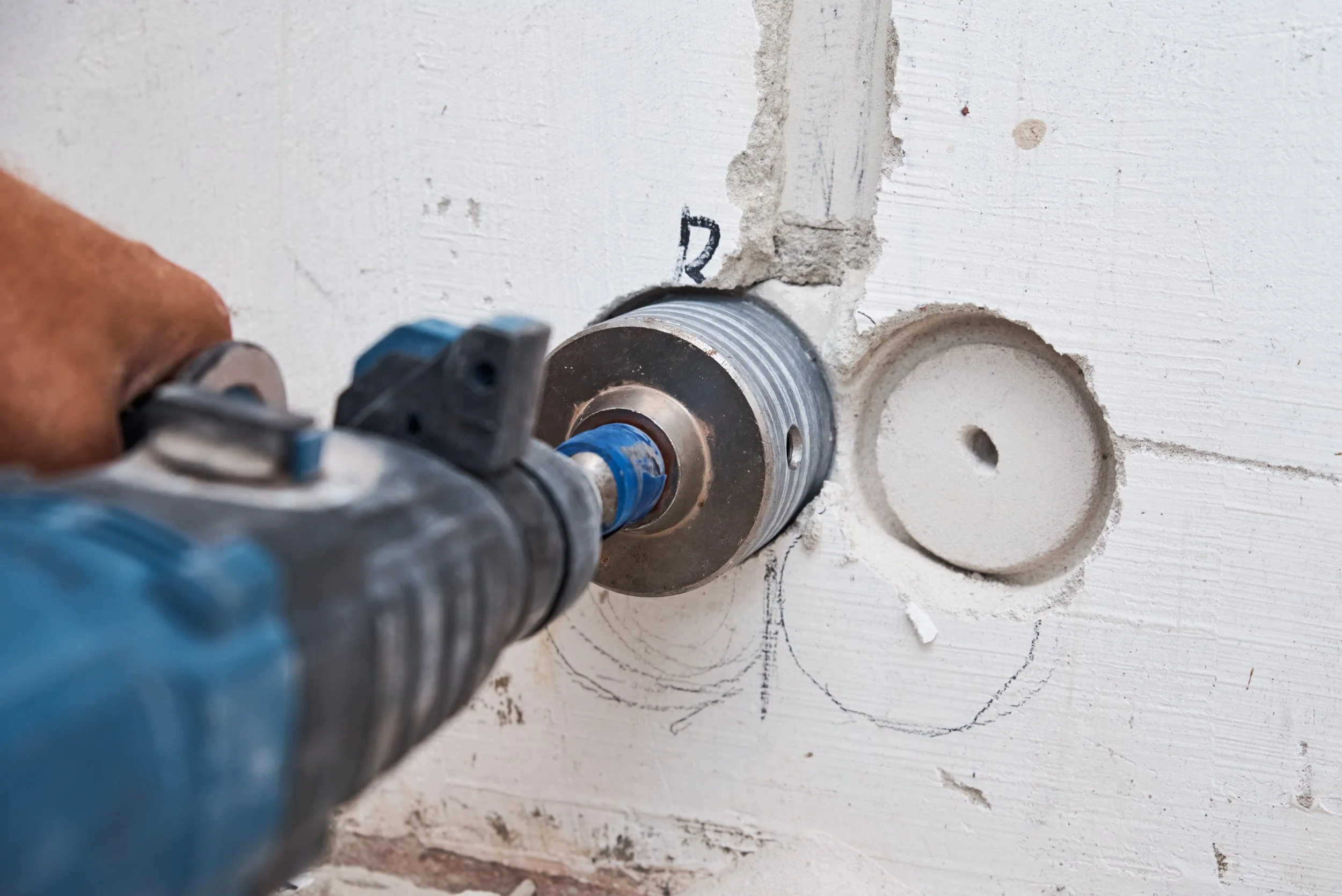A person using a power drill on a wall, installing a circular electrical box or outlet, with a marked circle and lines around it for placement.