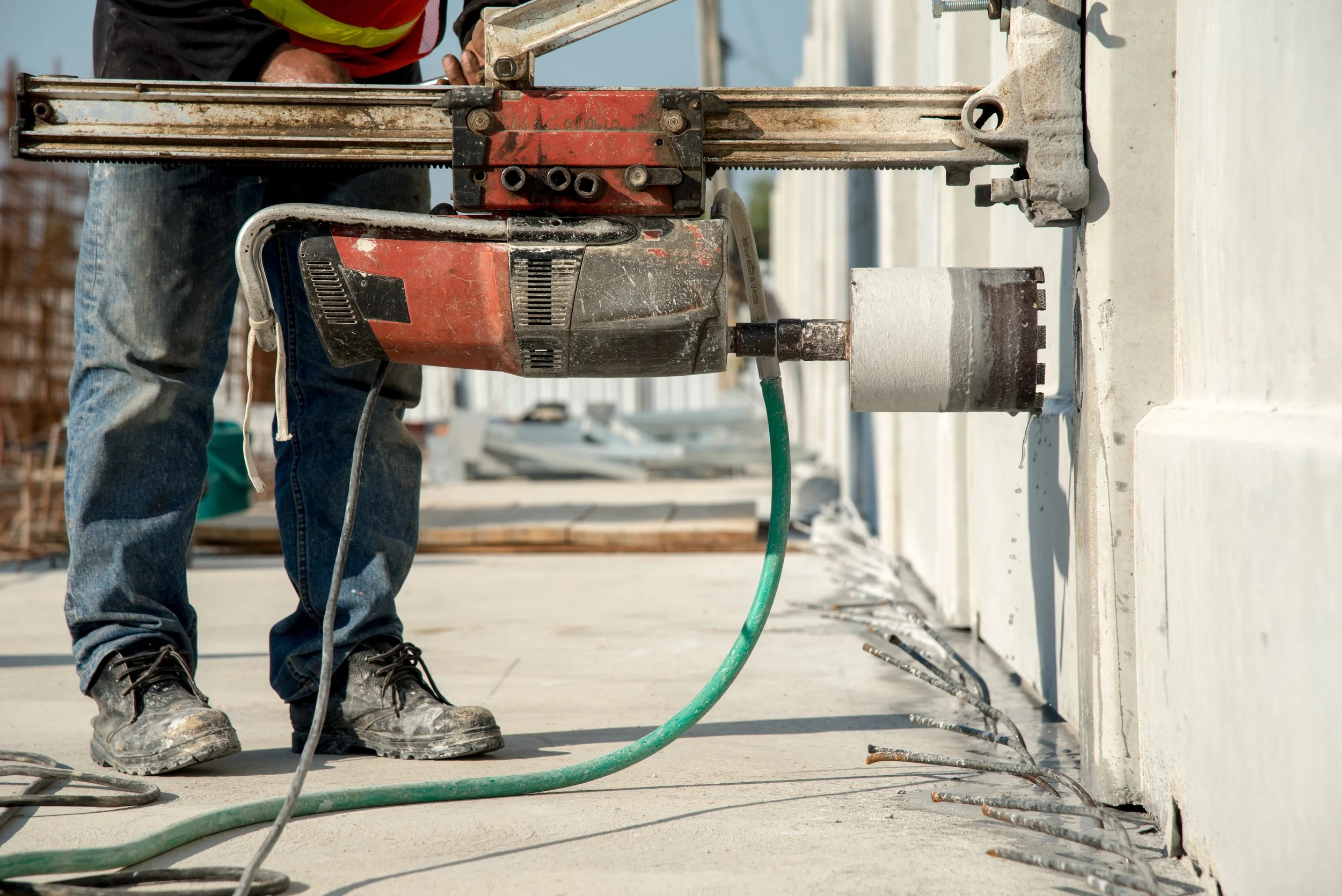 A worker at a construction site operating a large red and black power saw, cutting a concrete wall while wearing work boots and jeans.