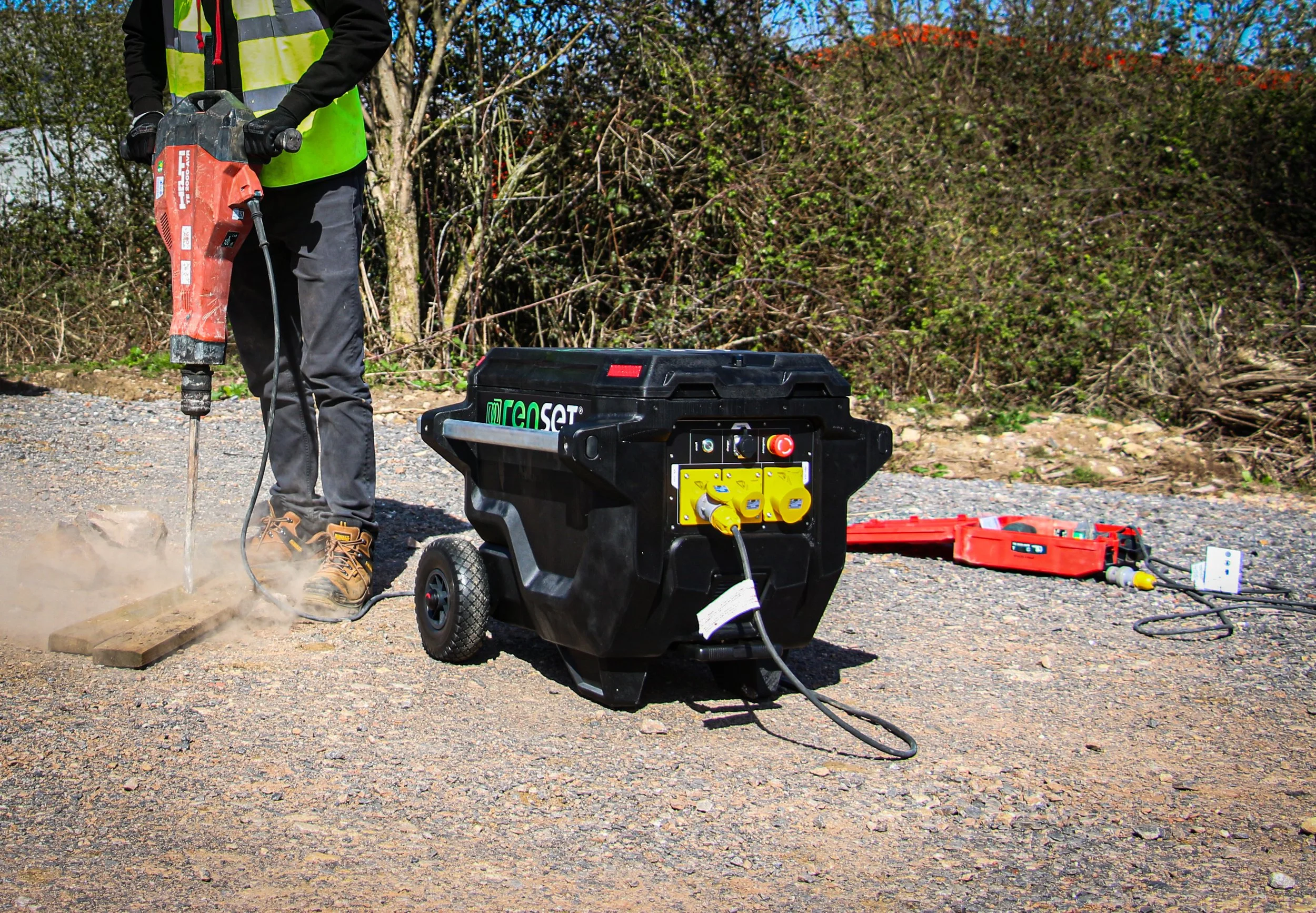 Person operating a large red and black power tool at an outdoor work site, with a black portable generator and open toolbox on the ground nearby, surrounded by trees and bushes.