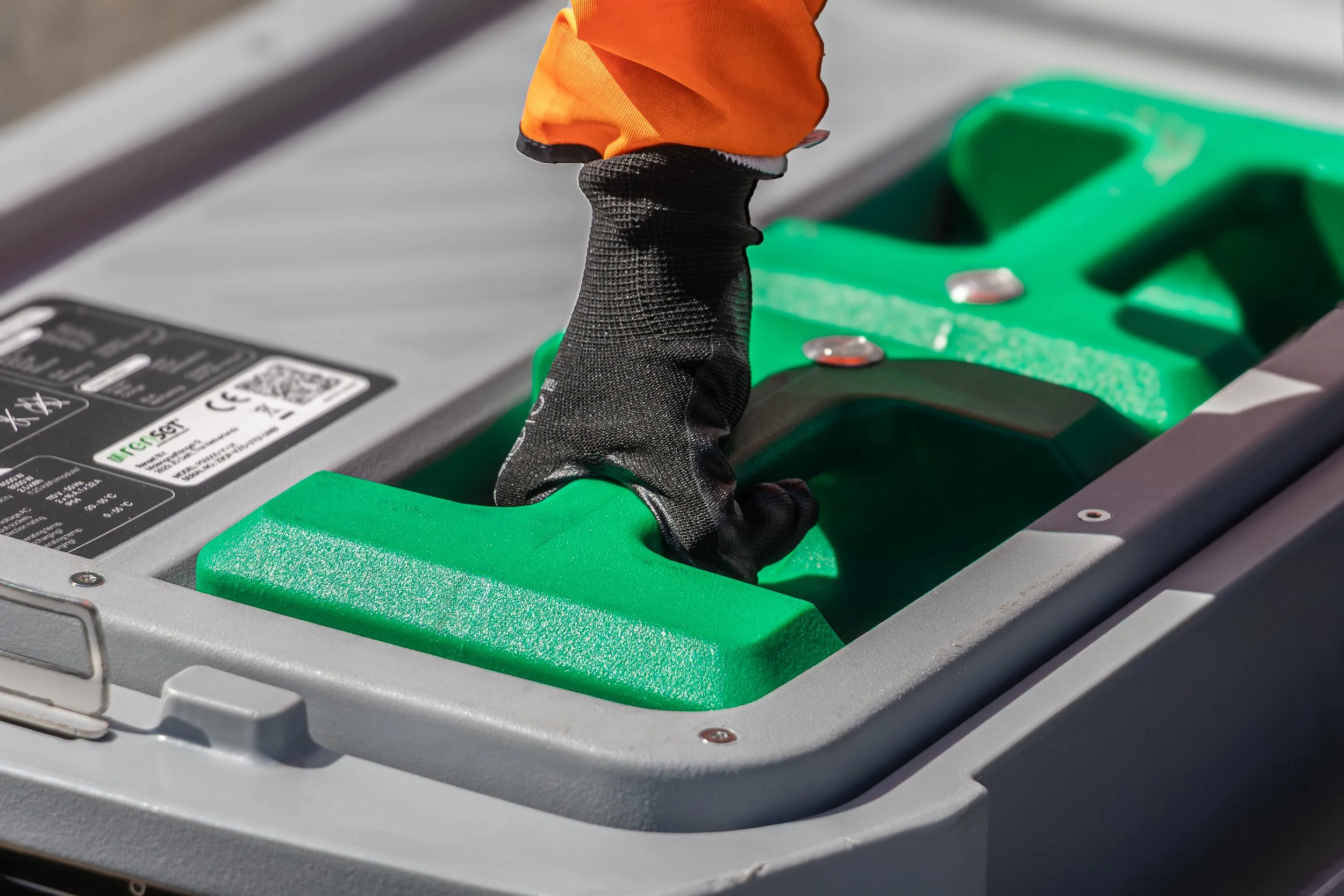 Close-up of a green electrical component or connector on a metallic surface with black textured tool or part-strap and person wearing orange sleeve nearby.