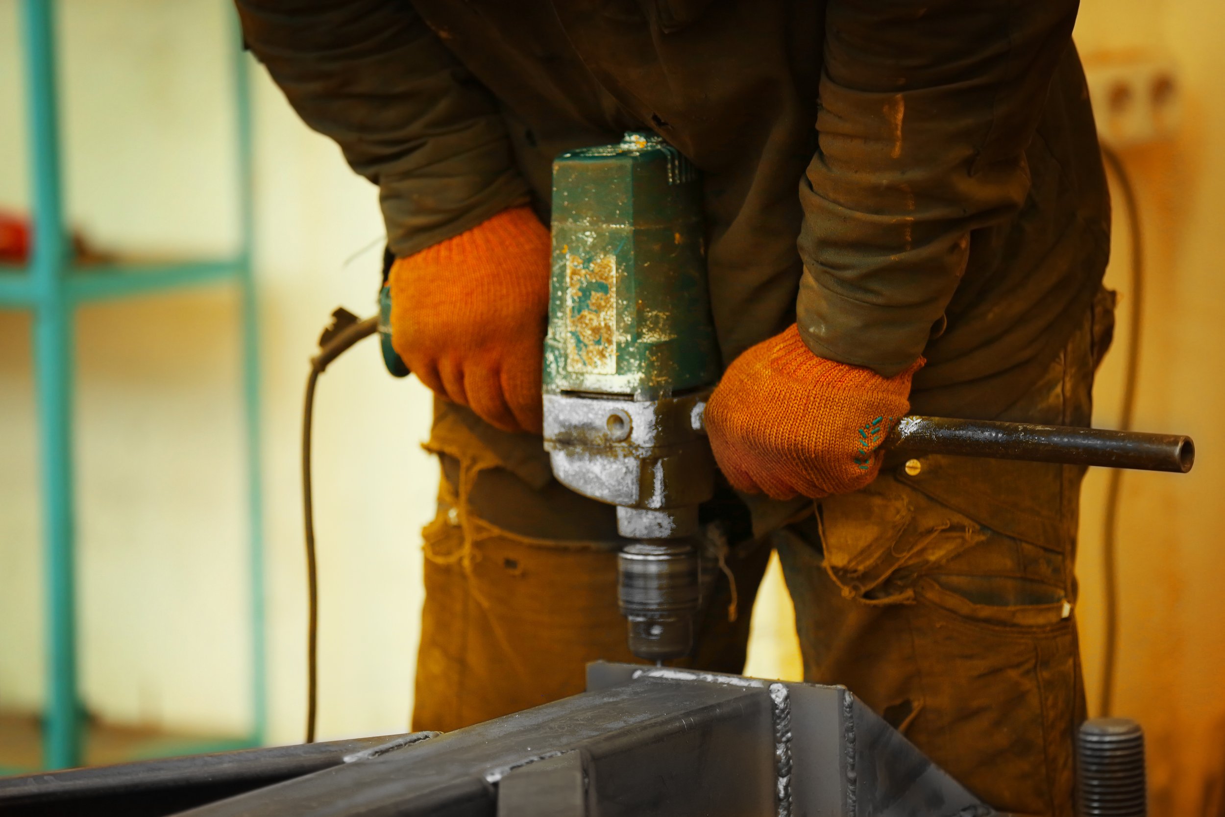 Person operating a power drill, wearing orange gloves and a dark work jacket inside a workshop.