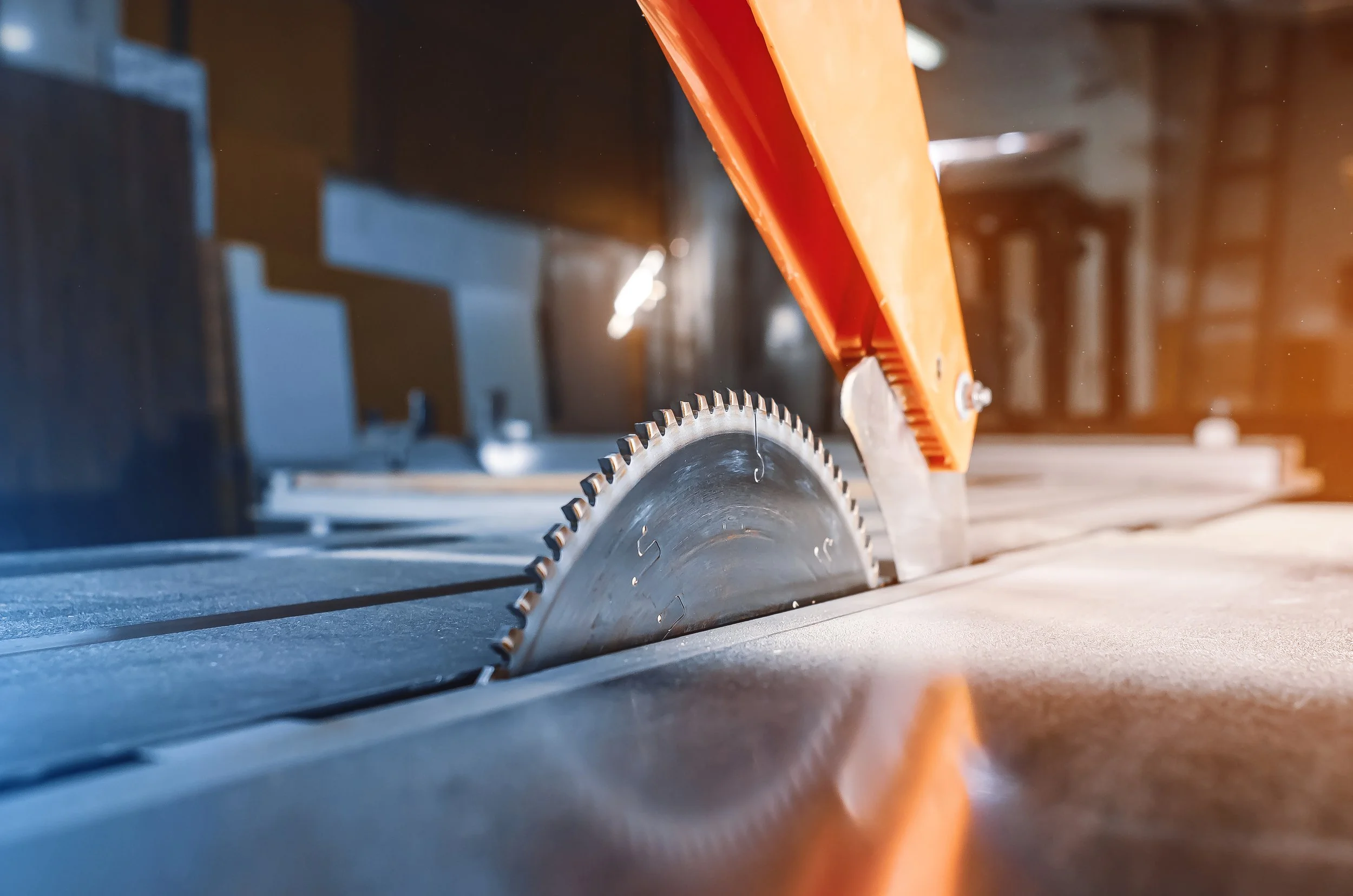 Close-up of a circular saw blade in a woodworking shop, with an orange safety guard above the blade.