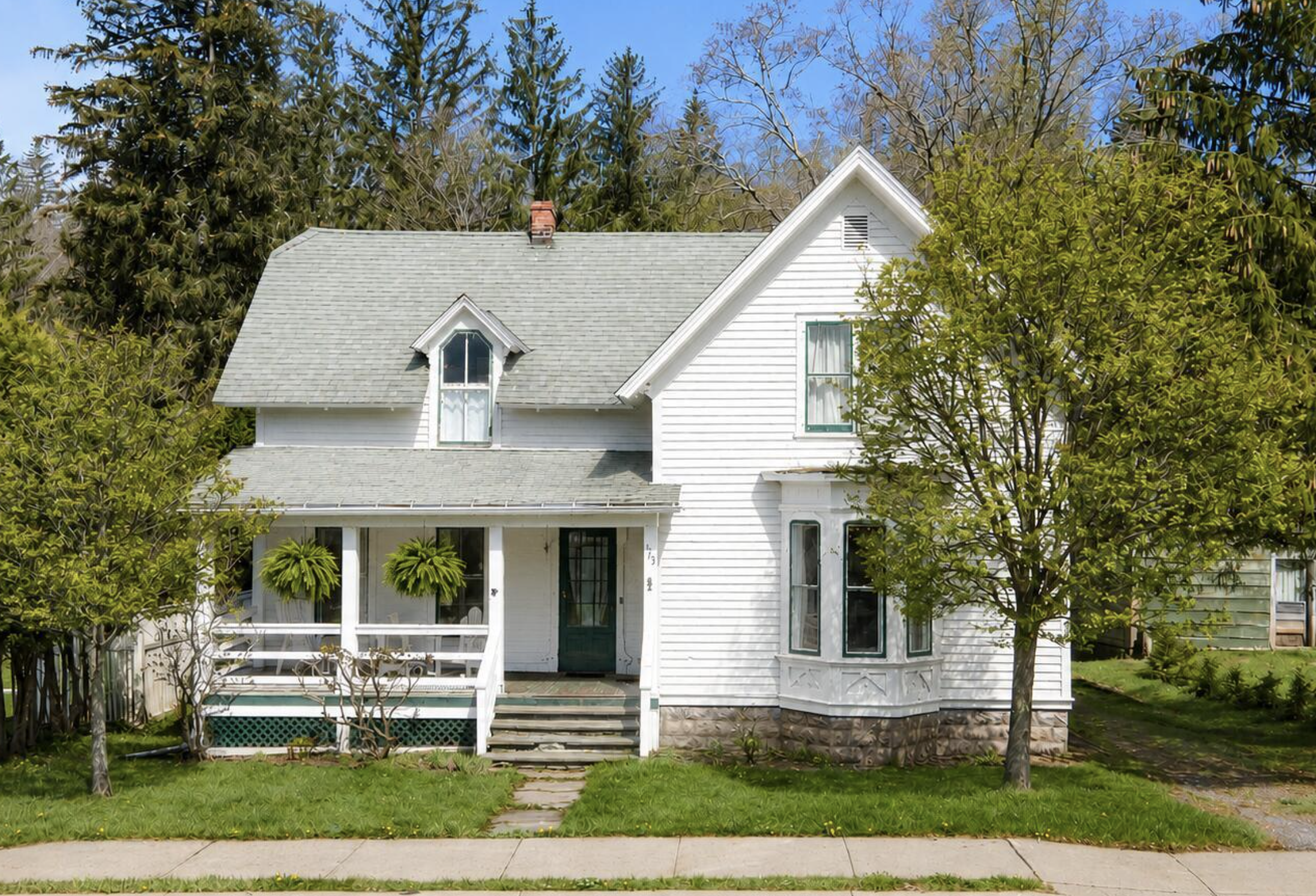 Front view of a white, two-story house with a porch and stairs leading up to the front door; trees and bushes surround the house, and a sidewalk runs in front.
