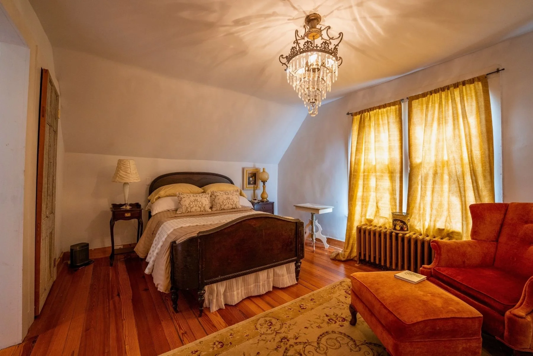 A cozy bedroom with a wooden bed, yellow curtains, an orange armchair with an ottoman, a chandelier hanging from the ceiling, and a white side table near the window.