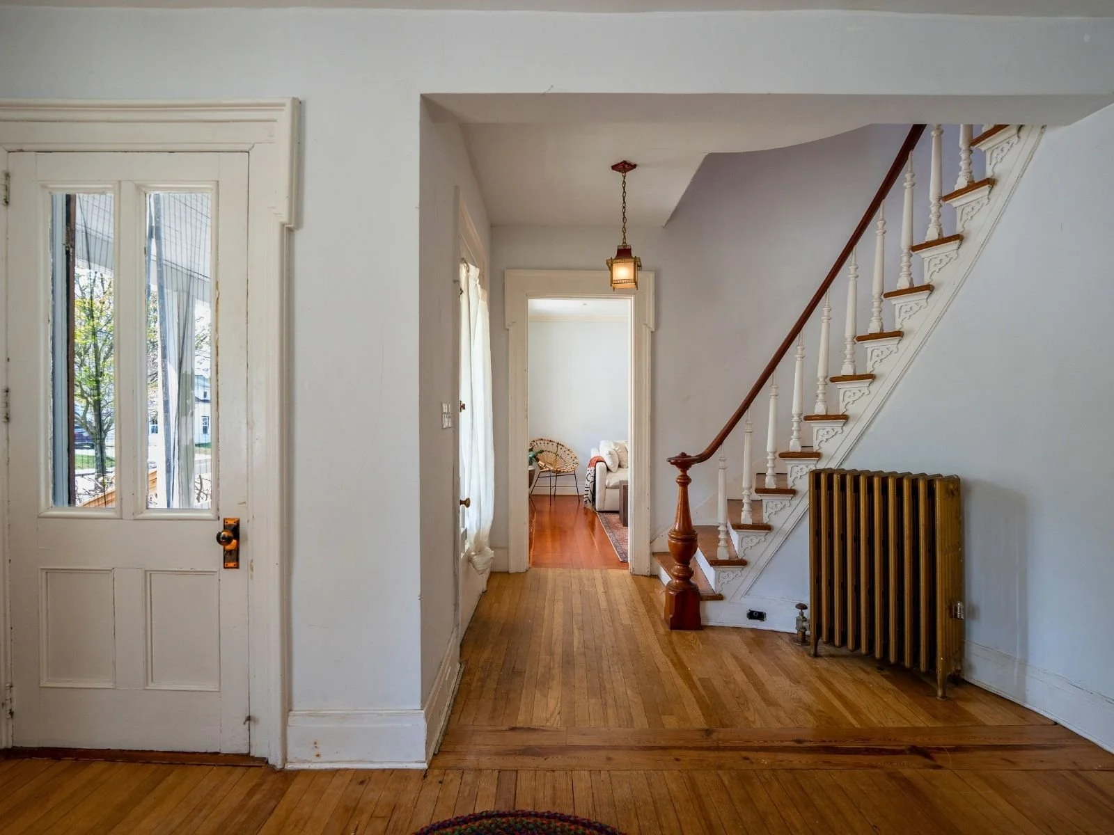 Interior of a house with a wooden floor, white walls, a stairway with a wooden banister on the right, a radiator, a doorway leading to a living room with a beige couch, and a door with glass panels on the left. There is a hanging light fixture in the hallway.