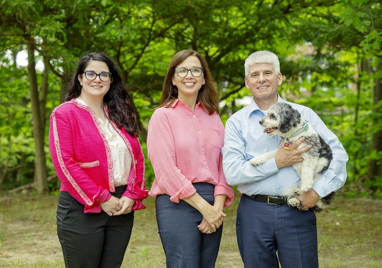 Three people and a dog in a park with green trees in the background. Two women and one man, all smiling, with the man holding a gray and white dog.