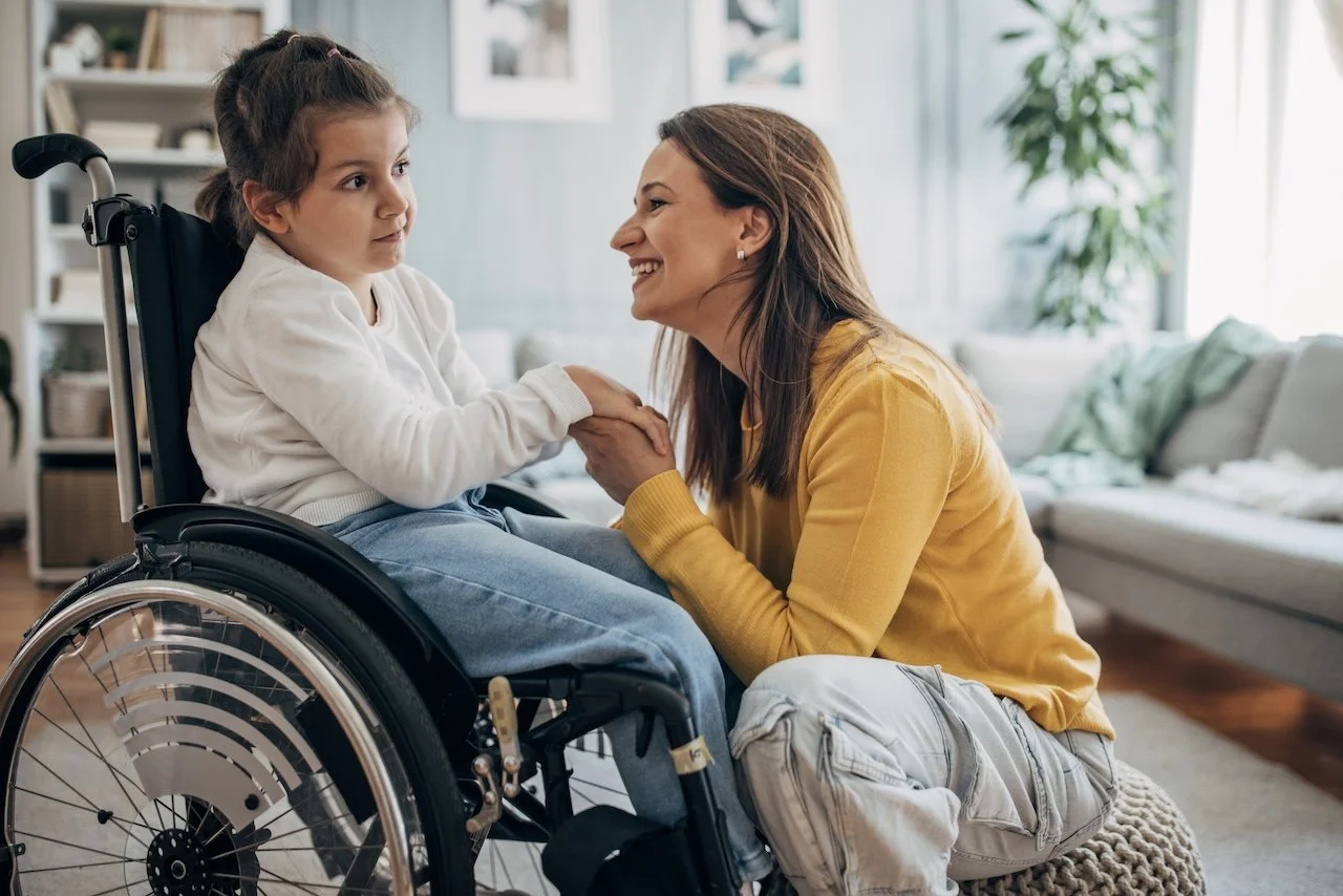 A woman smiling and holding hands with a young girl in a wheelchair in a living room.