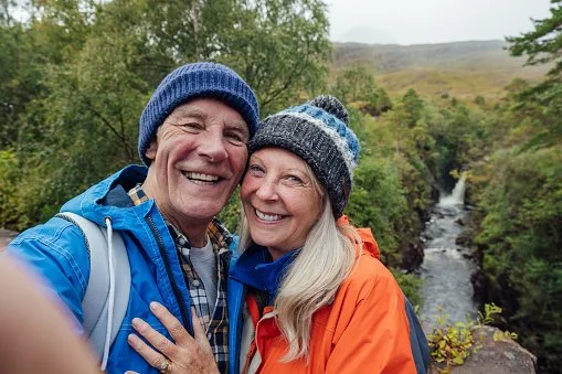 A smiling couple in outdoor gear taking a selfie with a waterfall and trees in the background.