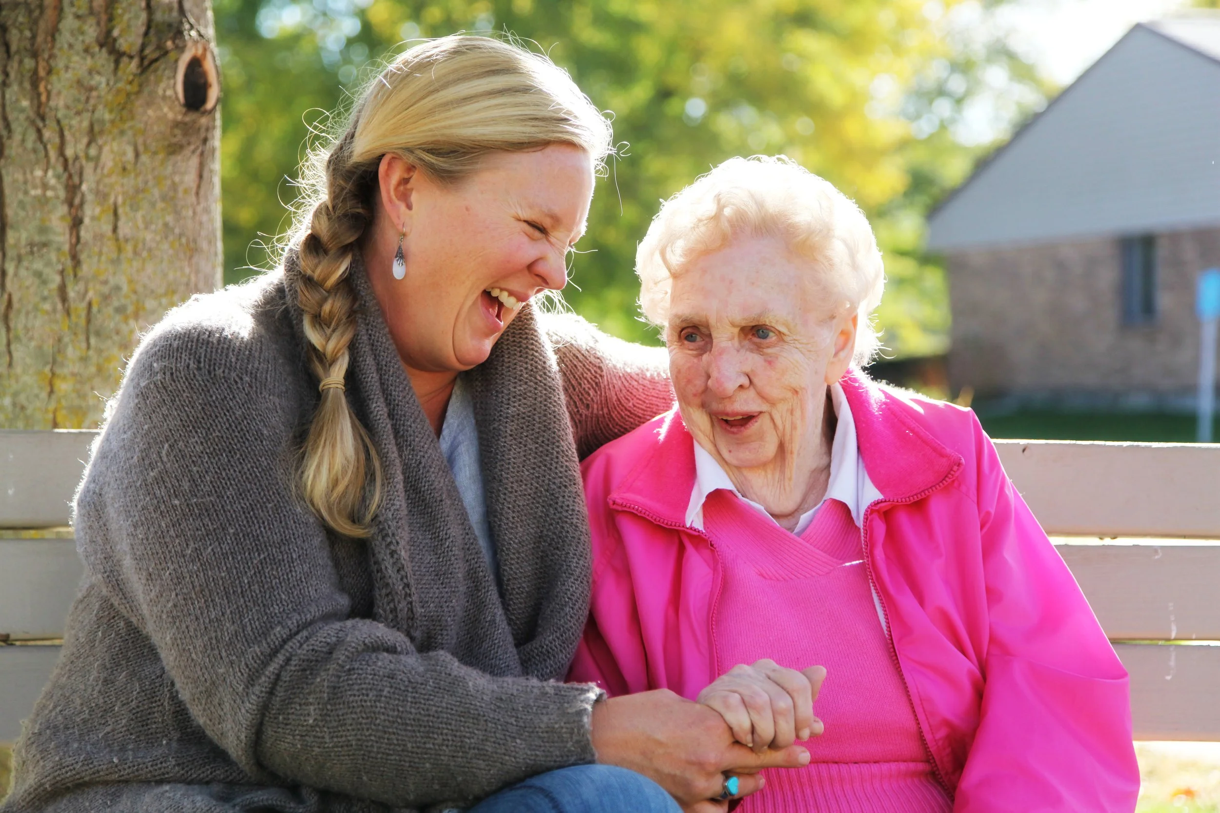 A woman with blonde hair in a braid and a woman with white hair sitting on a park bench, laughing and holding hands. They are outdoors with trees and a house in the background.