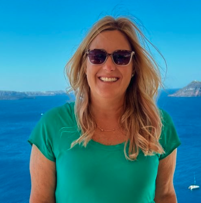 A woman smiling in sunglasses standing outdoors with a body of water and a boat in the background.