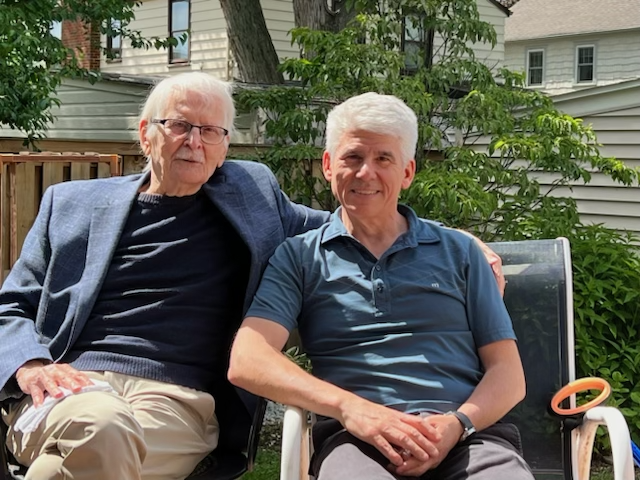 Two men sitting outside in a backyard, one older with glasses and white hair, wearing a gray blazer and black shirt, and the other middle-aged with gray hair, wearing a blue polo shirt. They are smiling and appear to be enjoying a sunny day near trees and houses.