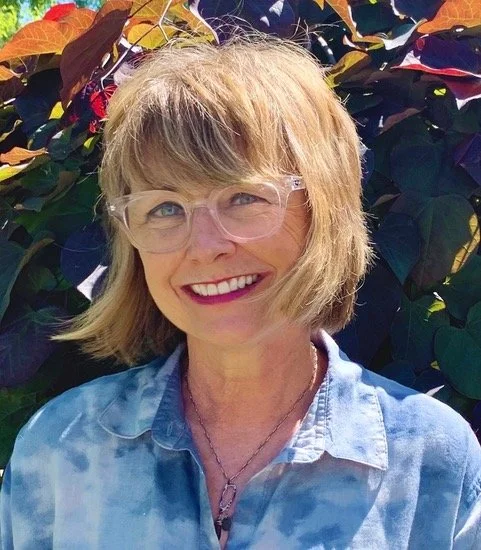 A woman with blonde hair, wearing glasses and a light blue shirt, smiling outdoors with colorful leafy plants in the background.