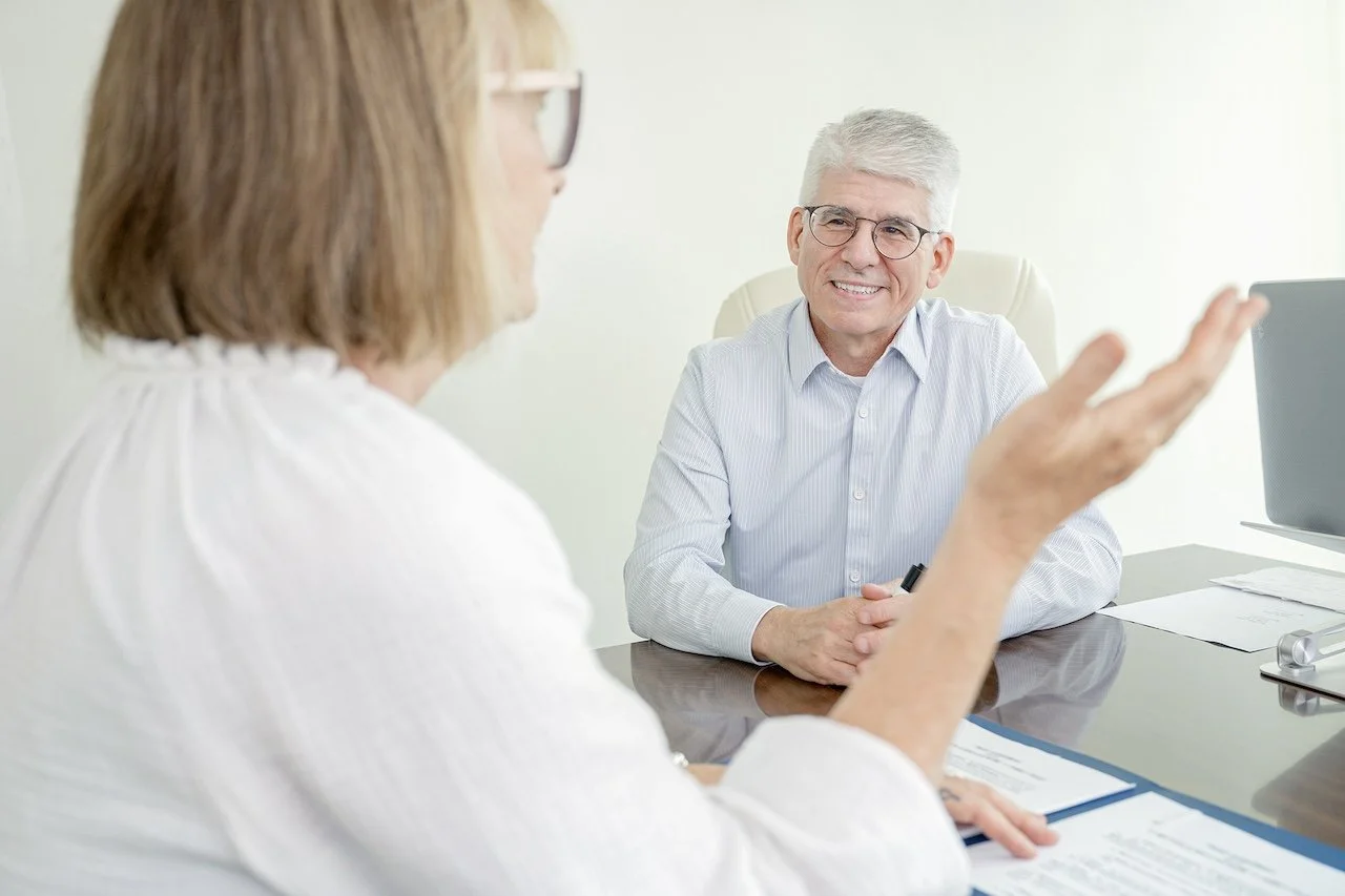 A man and a woman having a conversation in an office setting, with the woman gesturing with her hand and the man smiling.