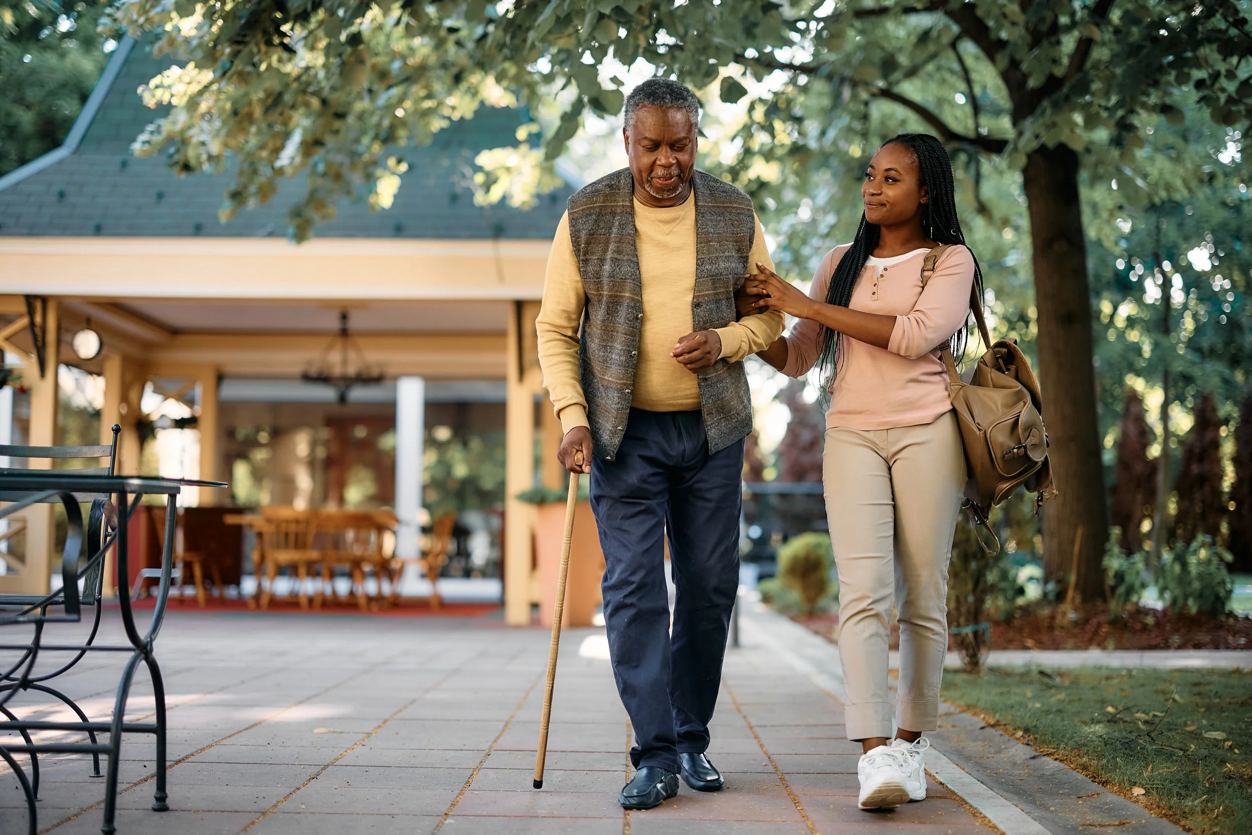 An elderly man with a cane walking outdoors with a young woman who is supporting him by holding his arm, in a park or garden setting with trees and a house in the background.