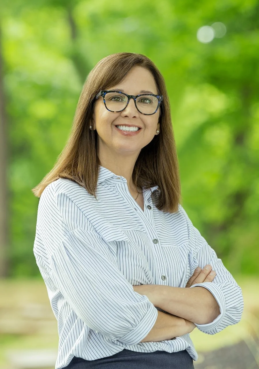 A woman with brown hair, glasses, and pearl earrings smiling with arms crossed outdoors with a green, leafy background.
