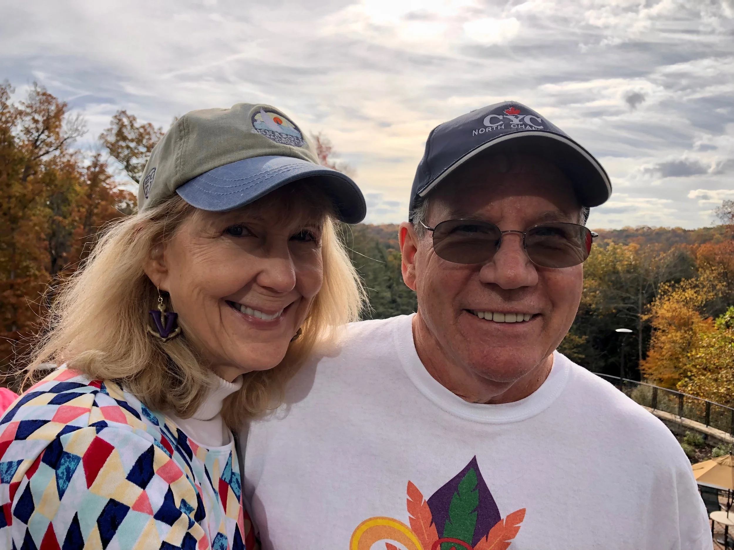 A smiling woman with blonde hair, wearing a multicolored geometric patterned shirt and a baseball cap, and a smiling man wearing glasses, a baseball cap, and a white T-shirt, standing outdoors during fall with colorful trees and overcast sky in the background.