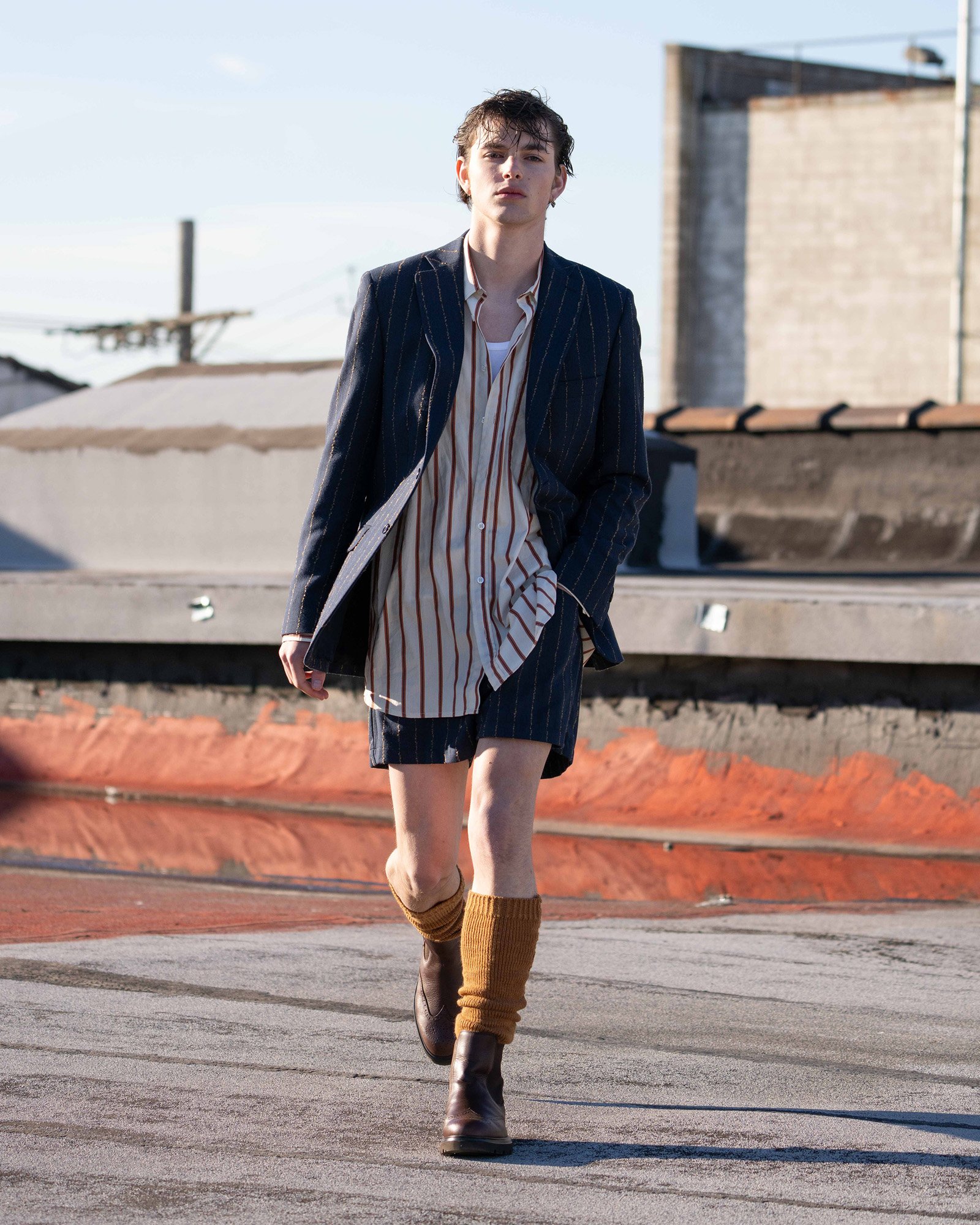 Person walking on a rooftop wearing a pinstripe suit with striped shorts, a long striped shirt, tan leg warmers, and brown boots.