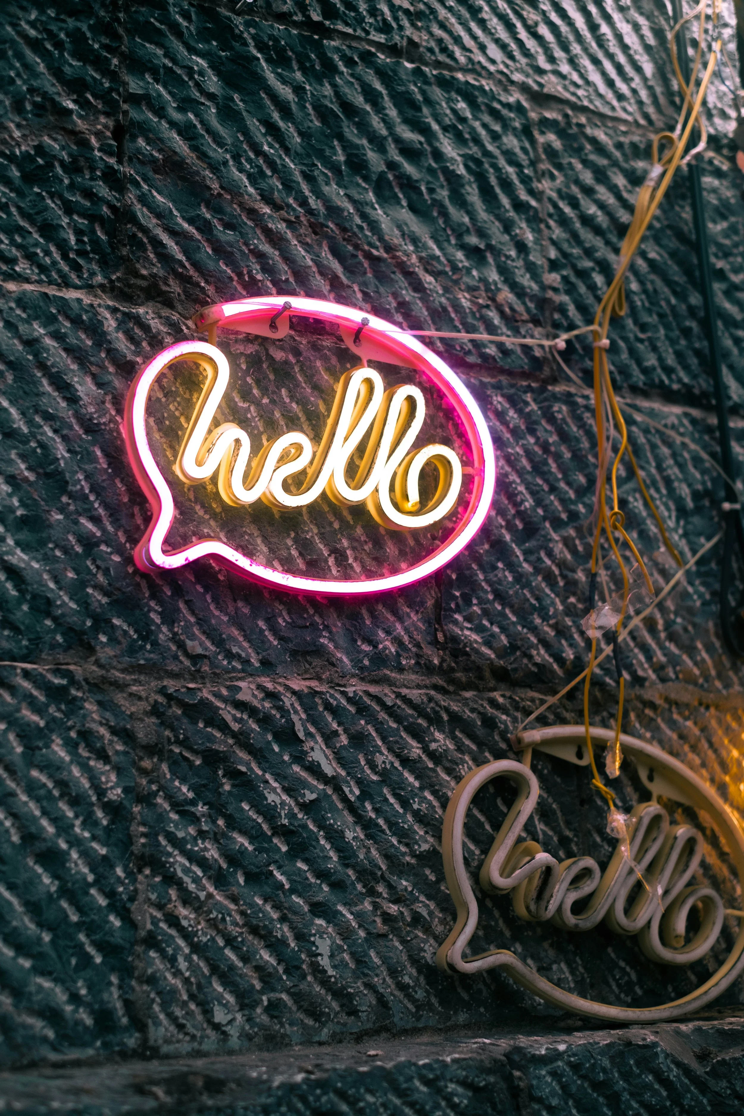 Neon sign on a brick wall with the word 'hello' inside a speech bubble outline.