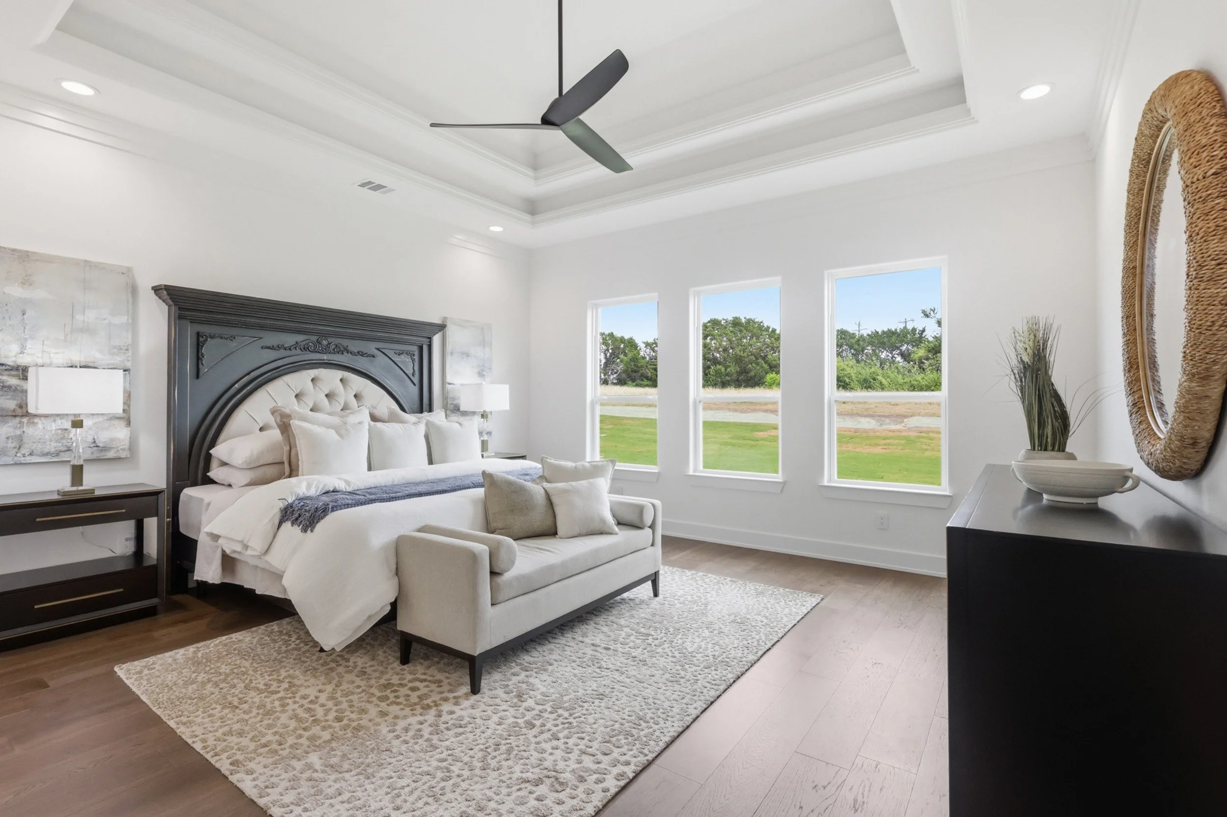 A modern bedroom with white walls and ceiling, ceiling fan, three windows showing trees outside, a bed with white bedding and a black headboard, two bedside tables with lamps, a beige armchair, and a beige patterned rug.
