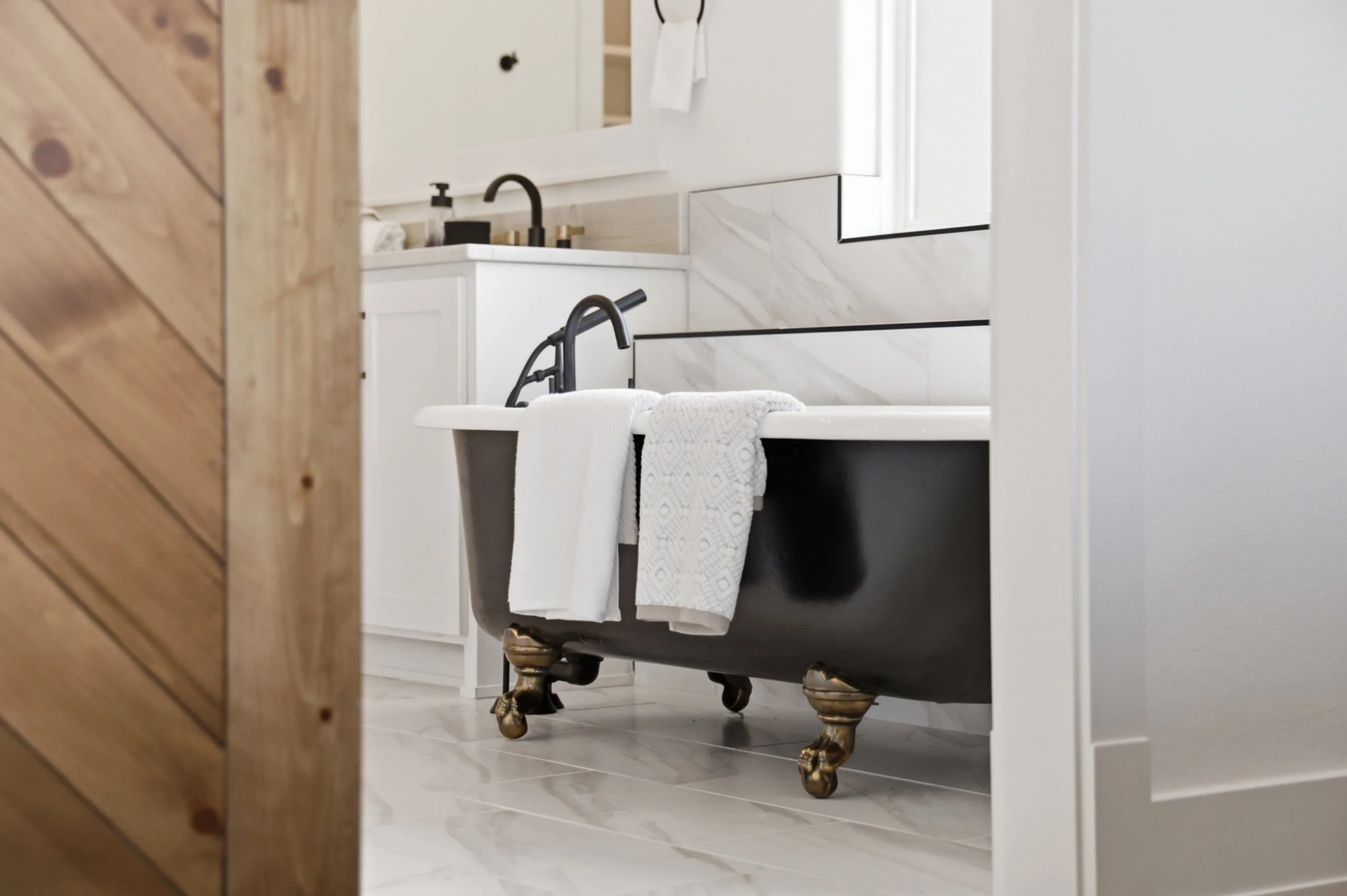 A vintage clawfoot bathtub with brass feet in a modern bathroom, partially visible through a doorway, with two white towels hanging on the bathtub.