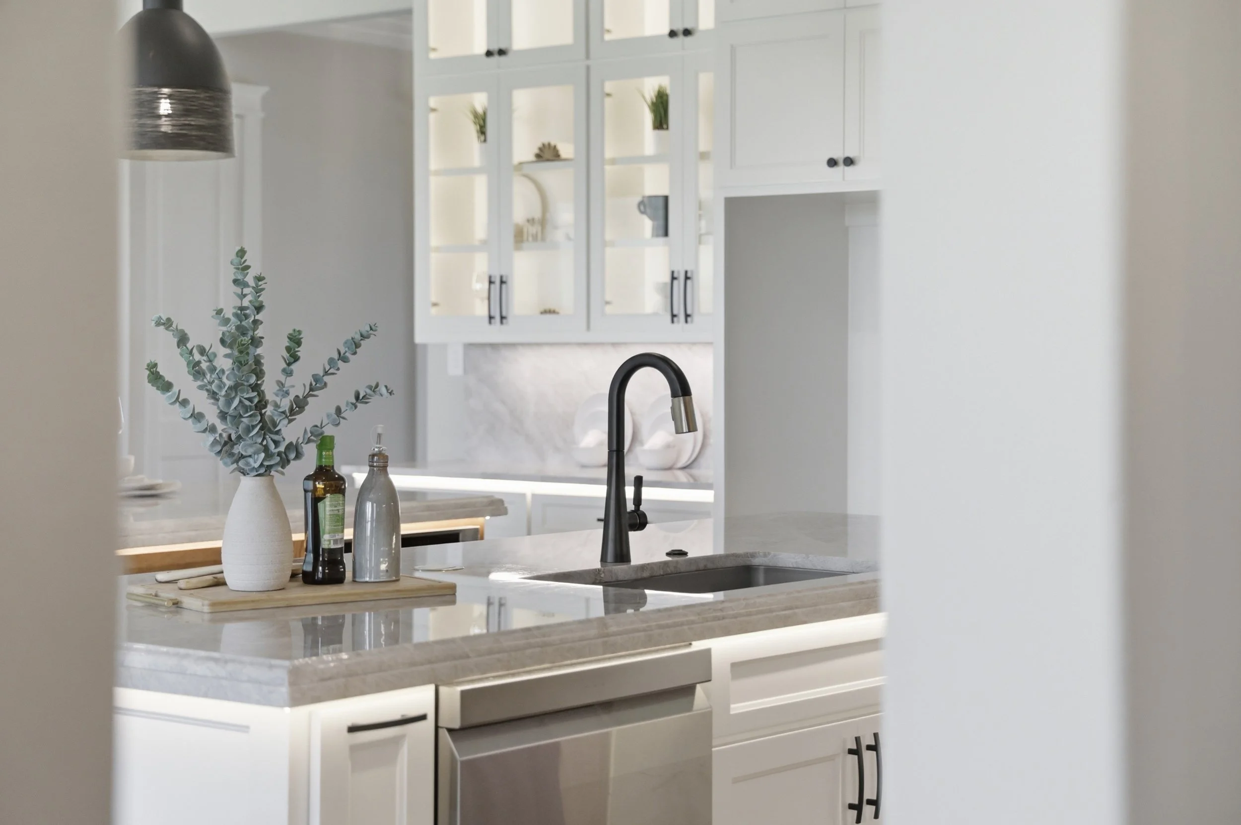 A modern kitchen with white cabinets, a beige marble countertop, and a black faucet. Decor includes a white vase with eucalyptus, bottled oils, and a cutting board. Sunlight enters the space, creating a bright atmosphere.