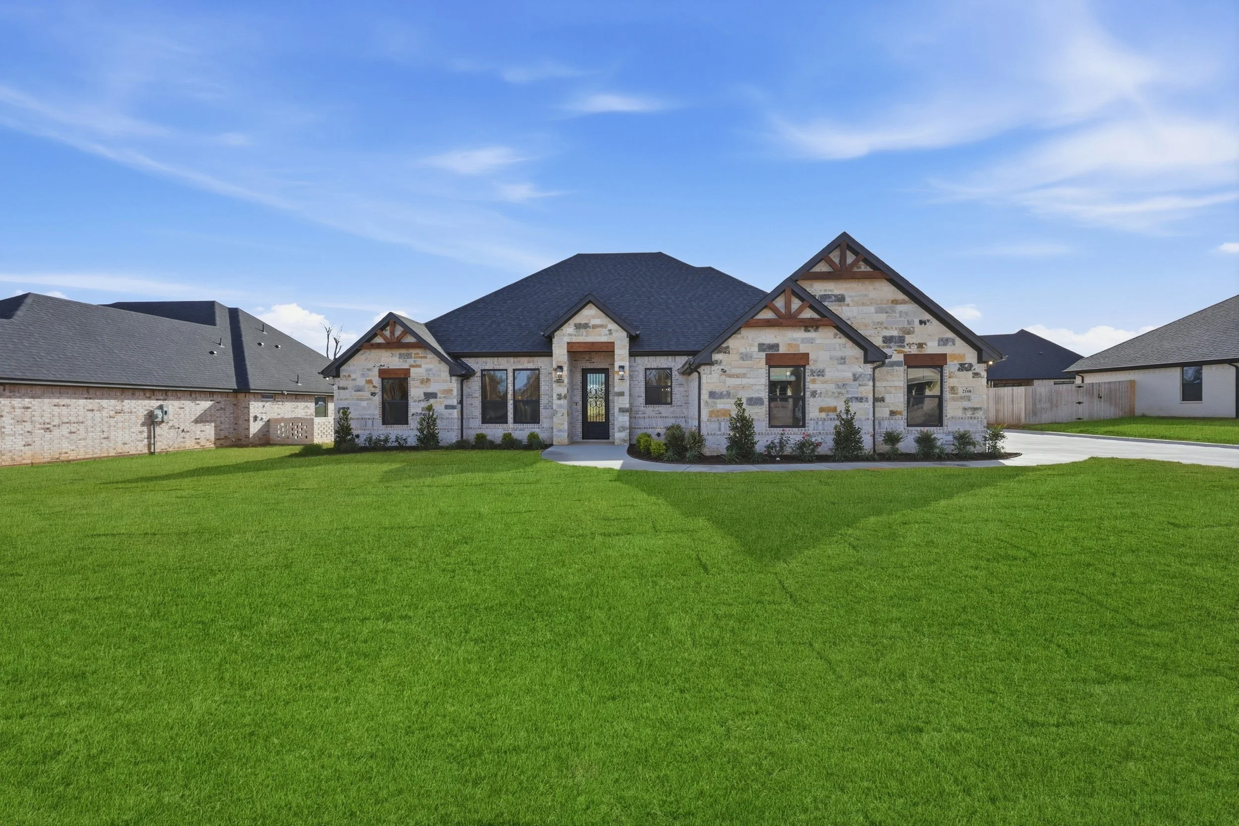 A modern house with a mostly brick exterior, large front windows, a black front door with glass, and a well-maintained grassy yard with small bushes. The house has a dark shingled roof and neighboring houses can be seen on either side. The sky is blue with some wispy clouds.