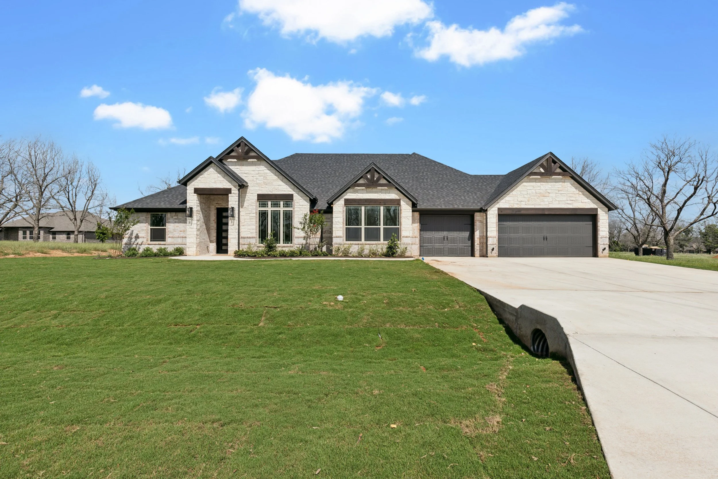 A modern, single-story house with stone and siding exterior, black shutters, and a gray roof. It has a two-car garage, a small front porch, and a well-maintained lawn with some small plants and trees. The sky is blue with a few clouds.