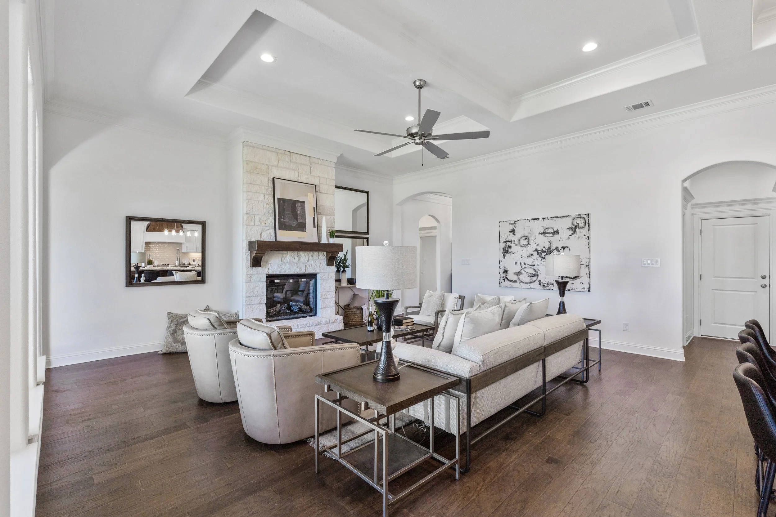 Living room with white walls, a stone fireplace, a ceiling fan, and plush seating including a sofa and armchairs, with artwork and lamps.