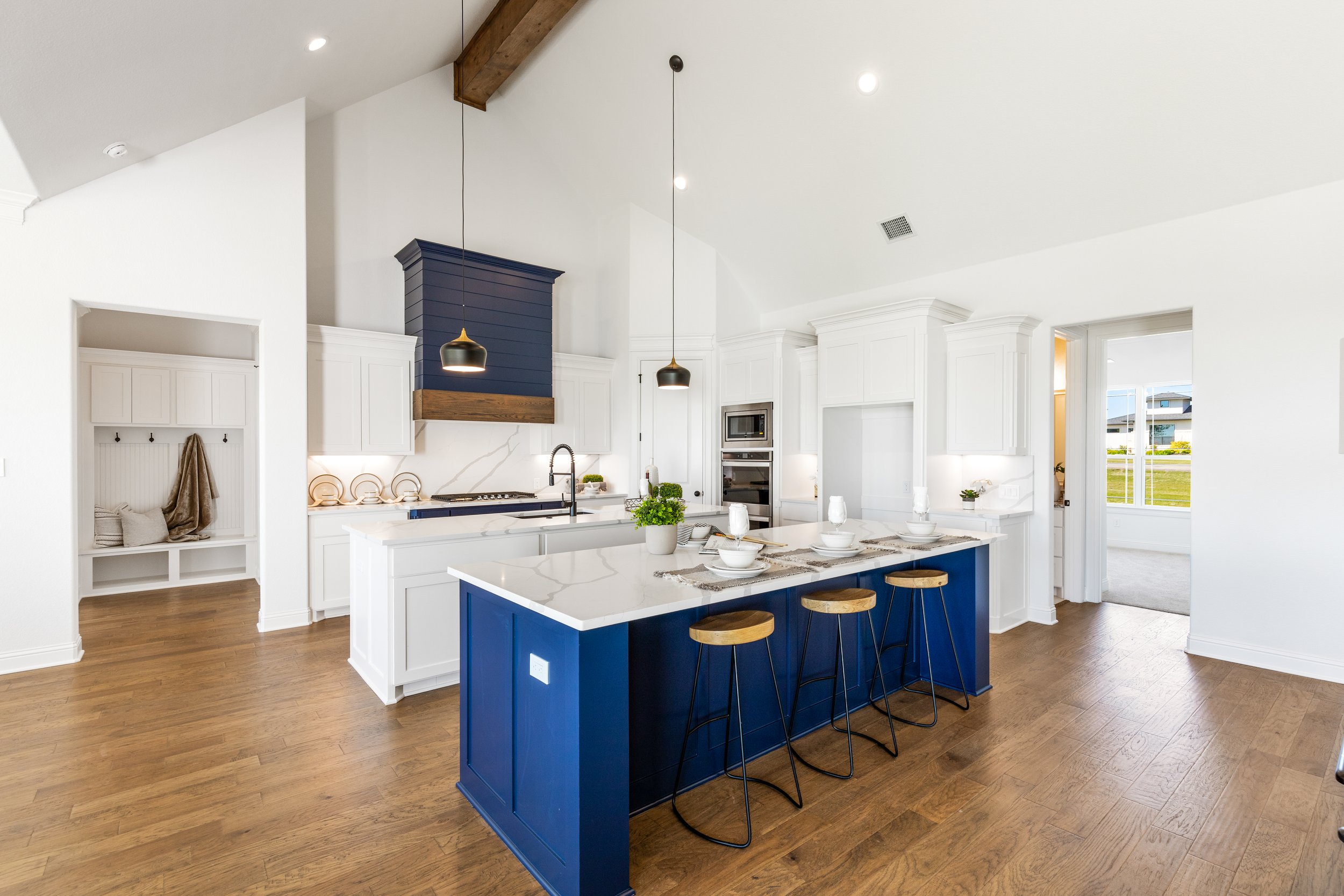 Modern kitchen with white cabinets, a blue island with a white countertop, and wooden bar stools. Pendant lights hang above the island. There is a large window allowing natural light, and a doorway leading to another room.