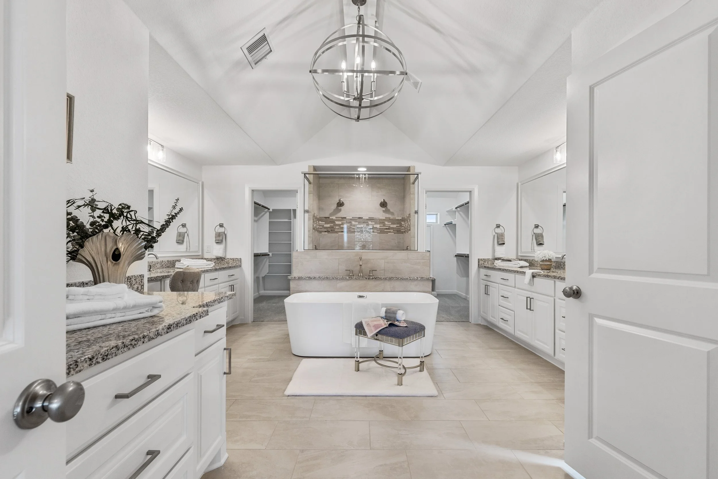 A modern, white bathroom with a large walk-in shower, a freestanding bathtub, double sinks, and ample storage. The space features sleek granite countertops, decorative towels, and a contemporary chandelier hanging from the ceiling.