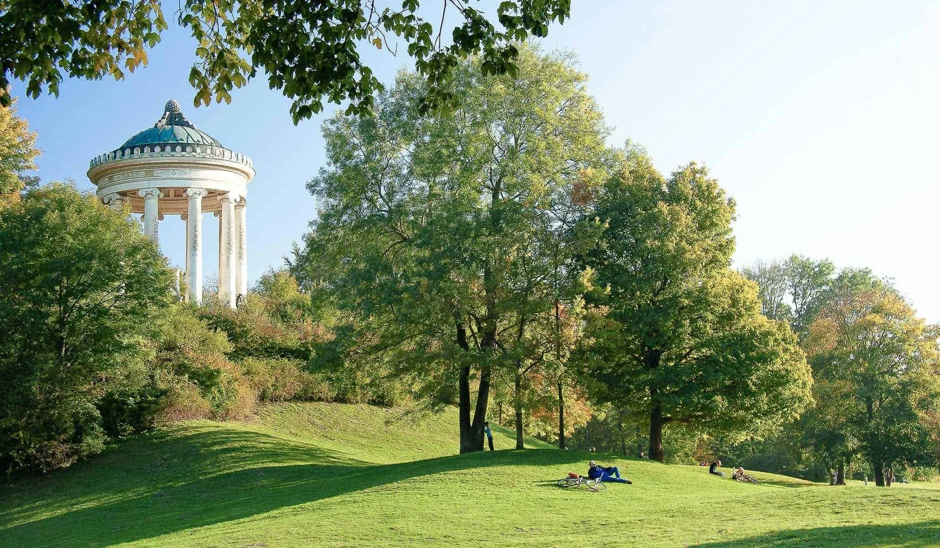 Parkanlage mit grünen Bäumen, einer Wiese und einer runden Tempelanlage mit Säulen im Hintergrund, einige Menschen entspannen auf der Wiese.