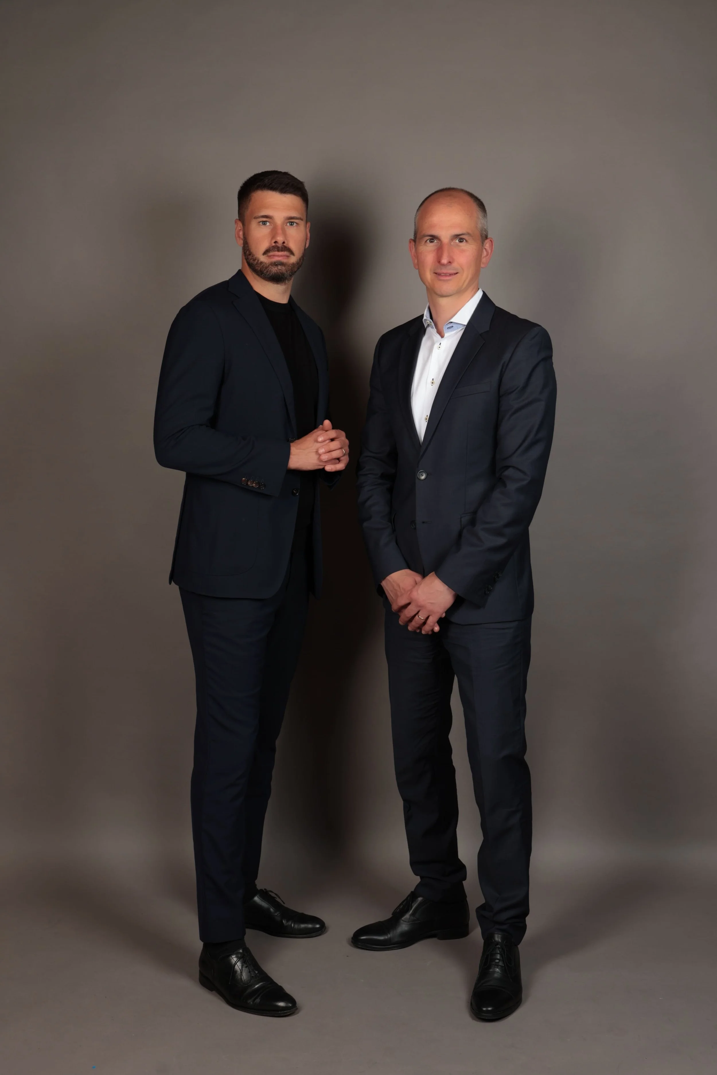 Zwei Männer in dunklen Anzügen stehen vor einem grauen Hintergrund.