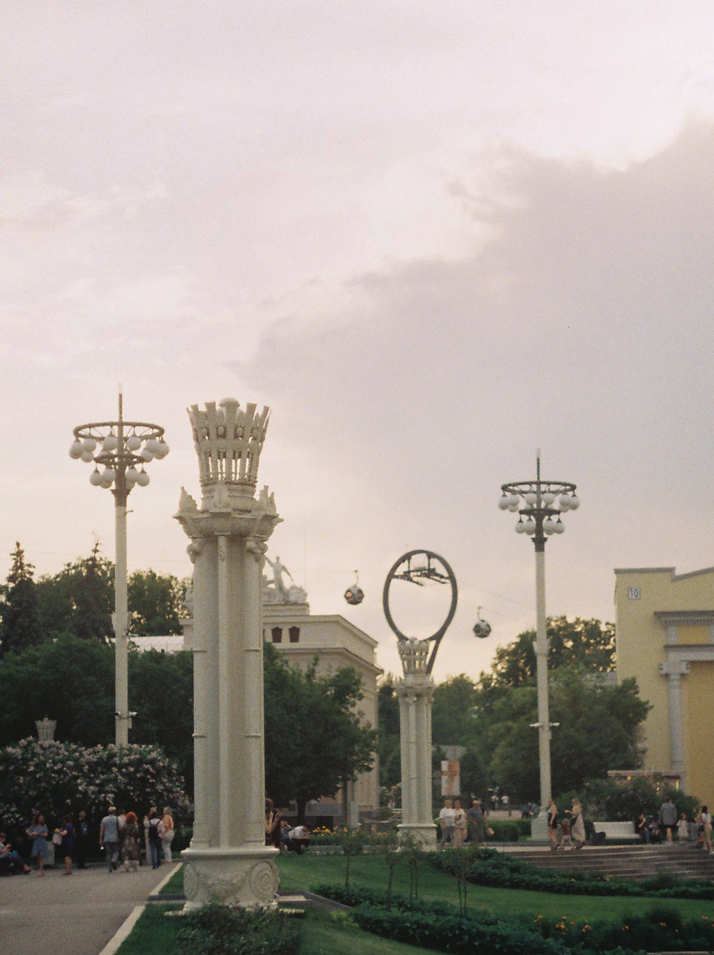 Public park with tall decorative lampposts, white columns with sculptures, and cable cars in the background on a cloudy day.