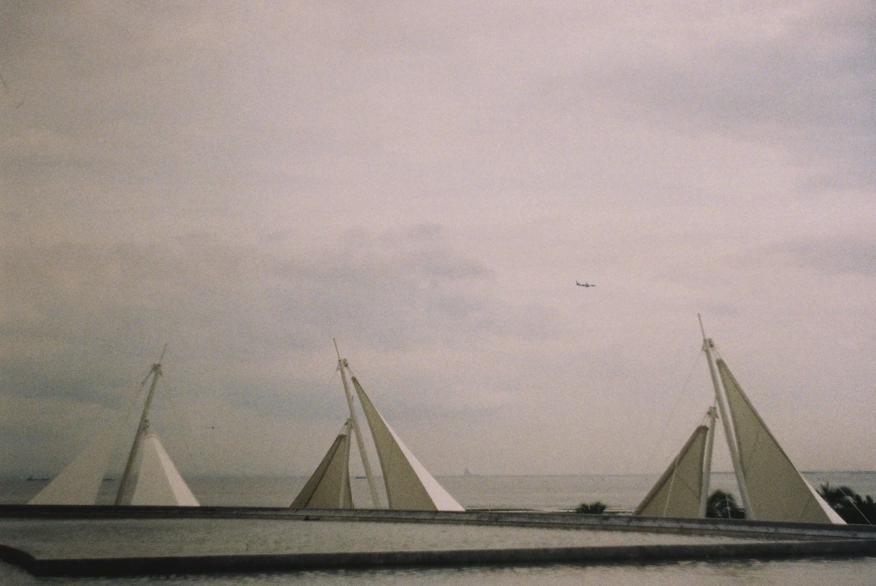 A cloudy sky with a small airplane flying above the top of a structure with white sails or tent-like covers supported by tall masts.