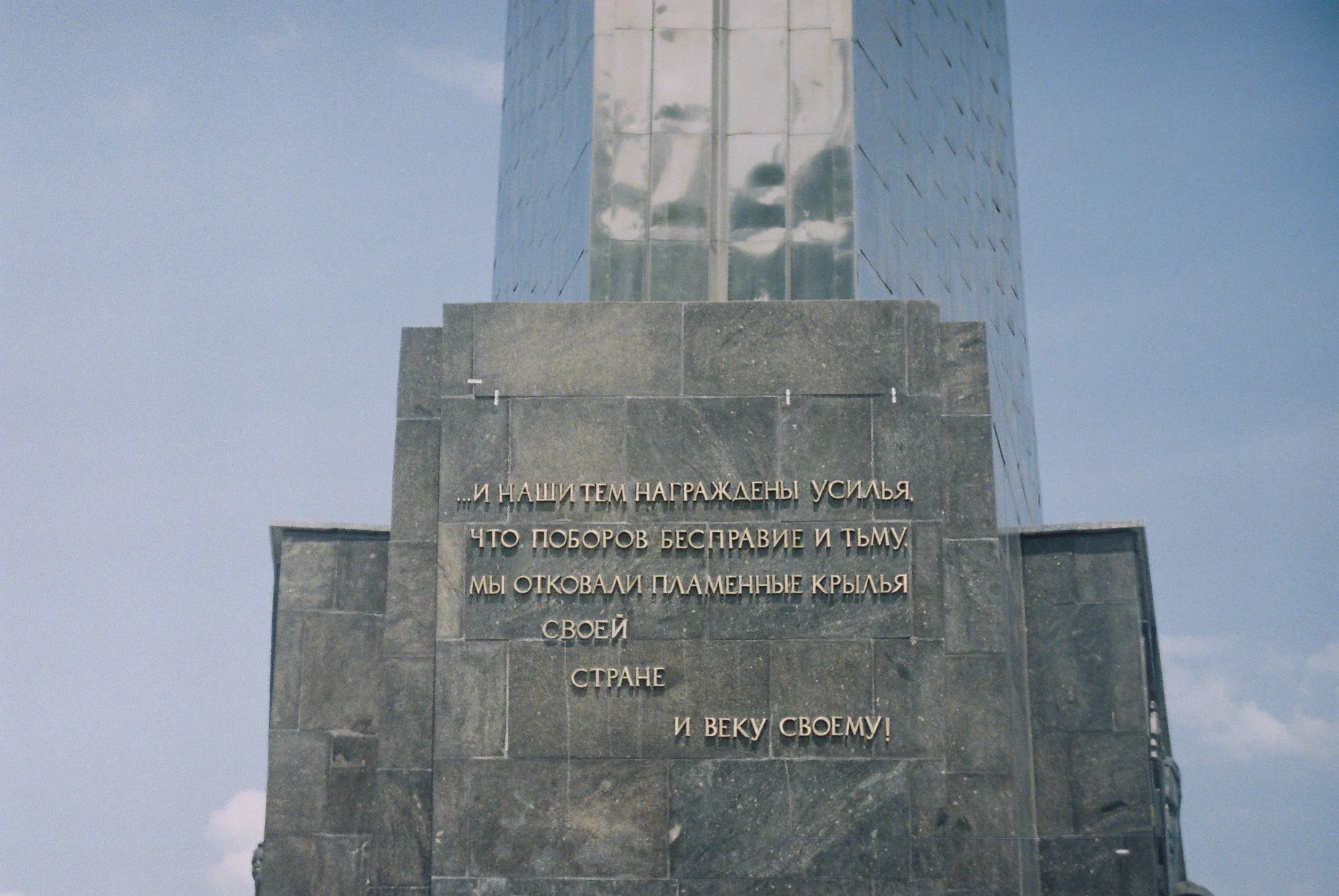 A tall monument made of dark gray stone with a reflective glass tower on top. The monument has an inscription in Russian on the front. The sky is partly cloudy.