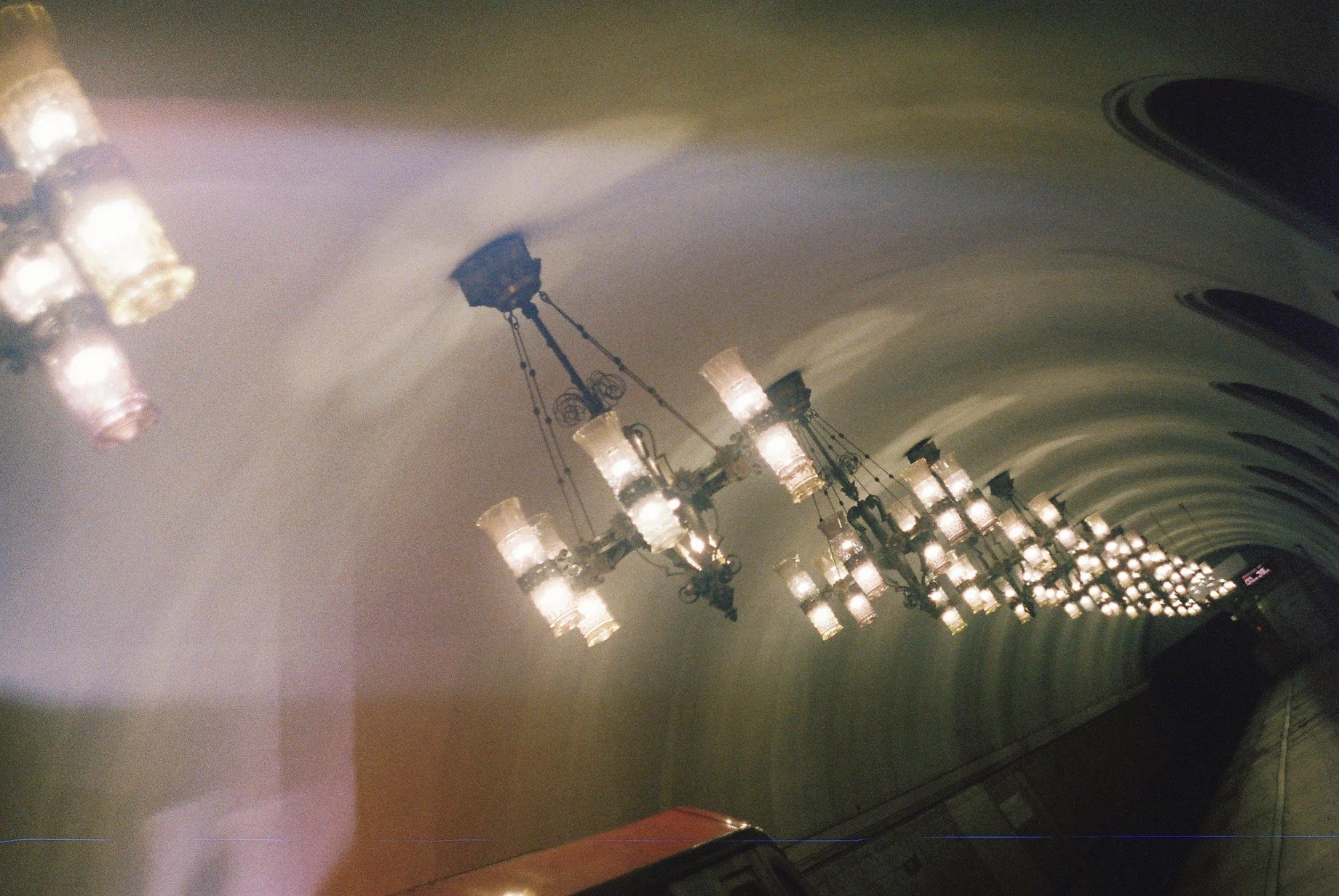 A long, arched hallway with a ceiling decorated with multiple chandeliers hanging from chains, illuminating the space with soft light.