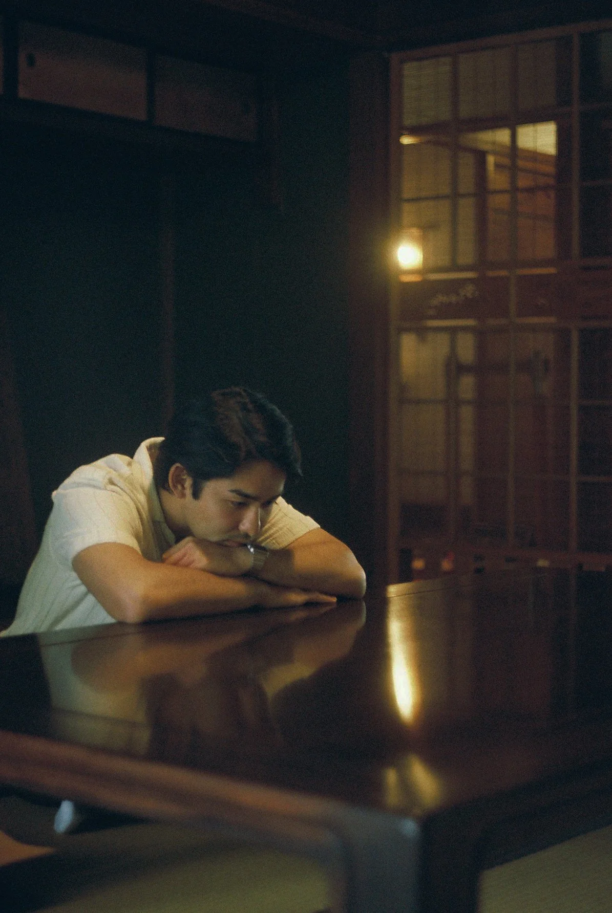A young man sitting at a dark wooden table, resting his head on his arms with a contemplative or sad expression in a dimly lit room with wooden lattice wall decor and soft lighting.