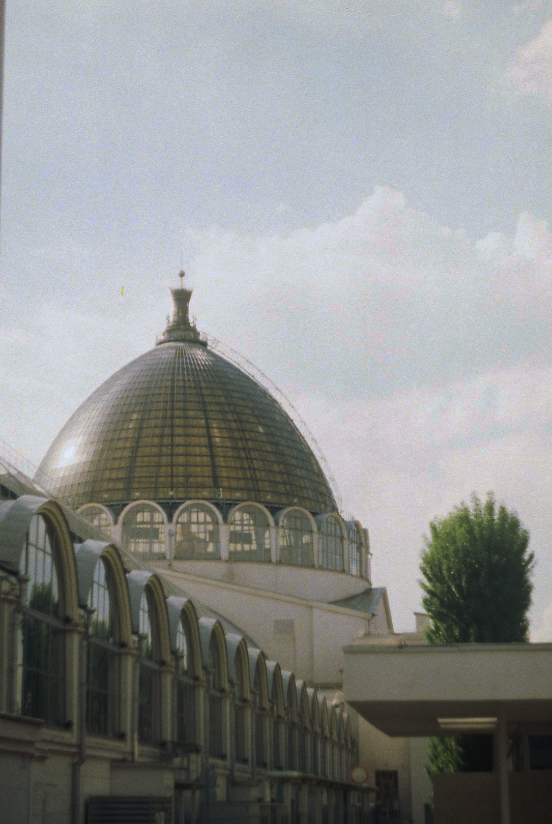 A large building with a prominent gold-domed roof and arched windows, with a second smaller building in front and greenery to the side, under a partly cloudy sky.