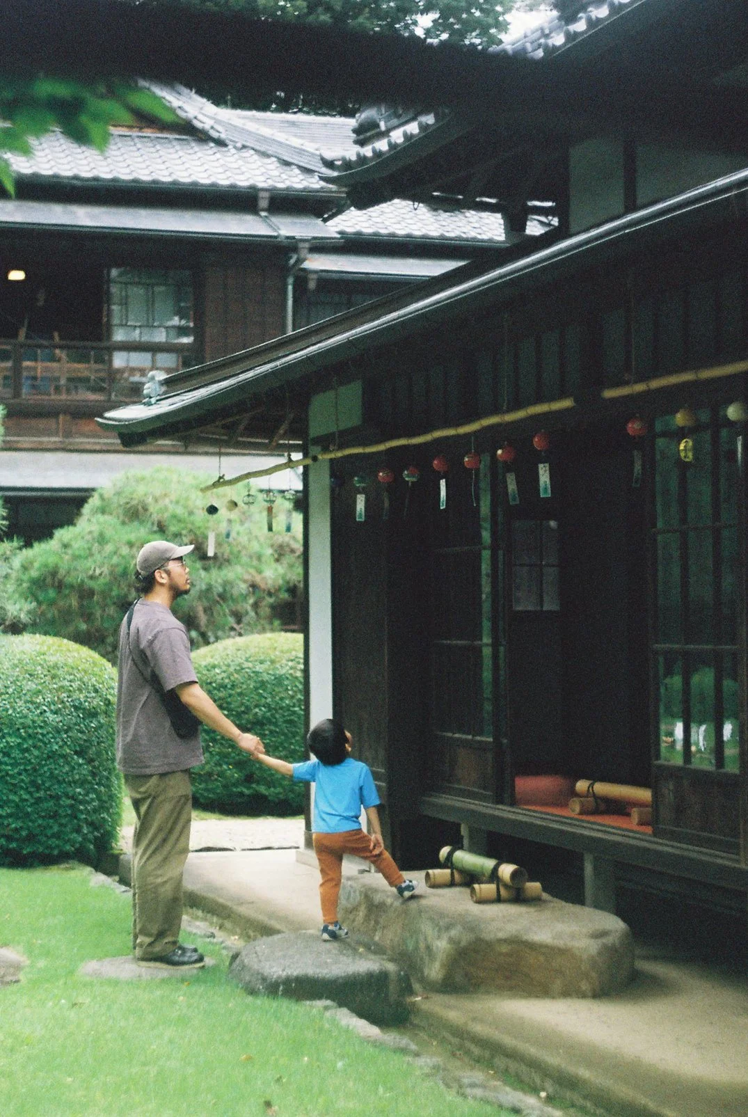 A man and a young child holding hands are standing outside a traditional Japanese building with dark wooden walls and tiled roof, near a lush green garden.