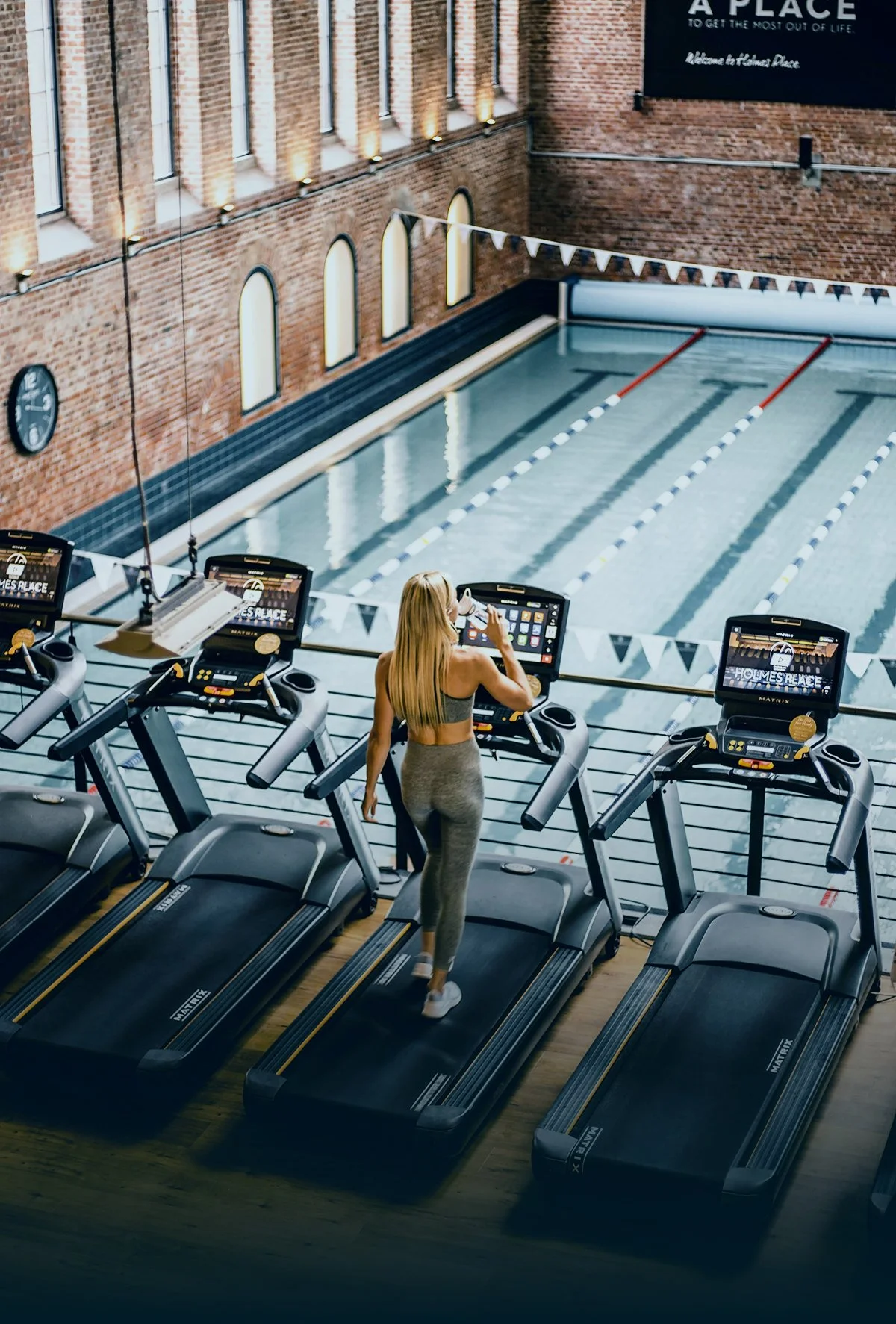Woman in workout clothes running on treadmill at indoor gym with pool in background and decorative brick wall.