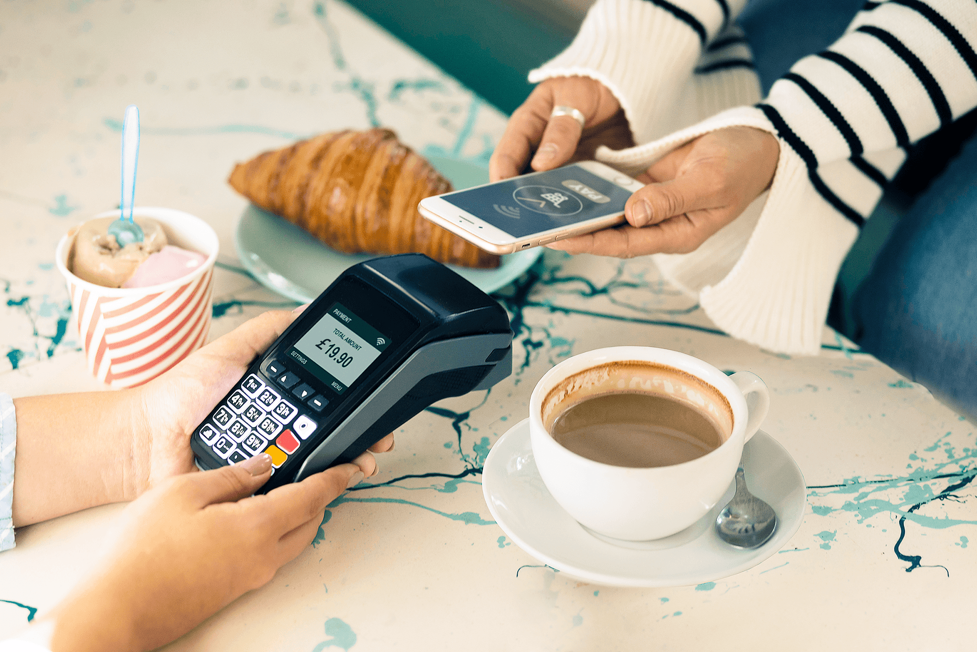 A person making a contactless payment via their phone and a handheld payment terminal next to some breakfast items.