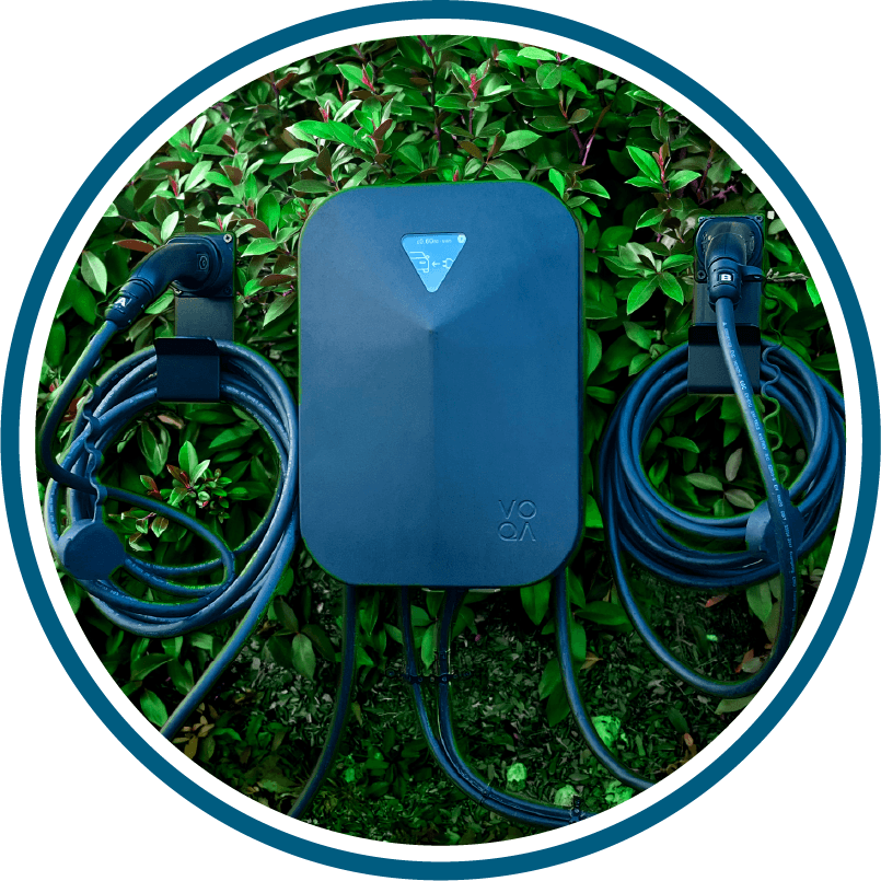 A Voqa electric vehicle charger with two tethered cables plugged into their holsters, mounted on a green bushy wall.